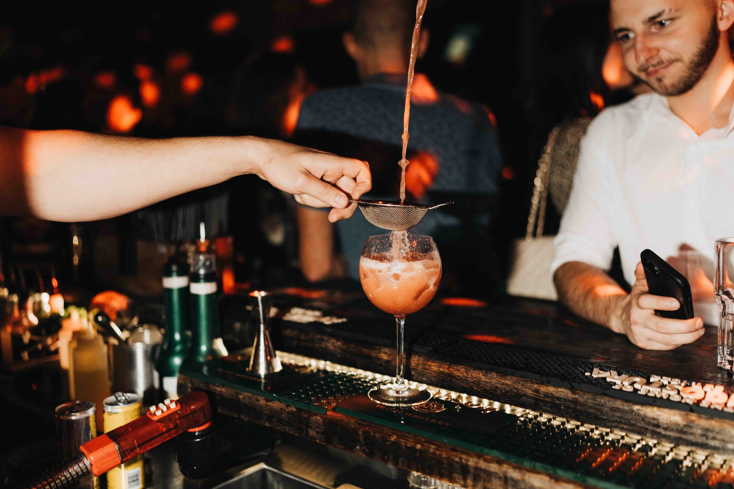 Bartender pouring a pink cocktail through a strainer into a wine glass at a dimly lit bar, with a man in a white shirt watching and using a smartphone.