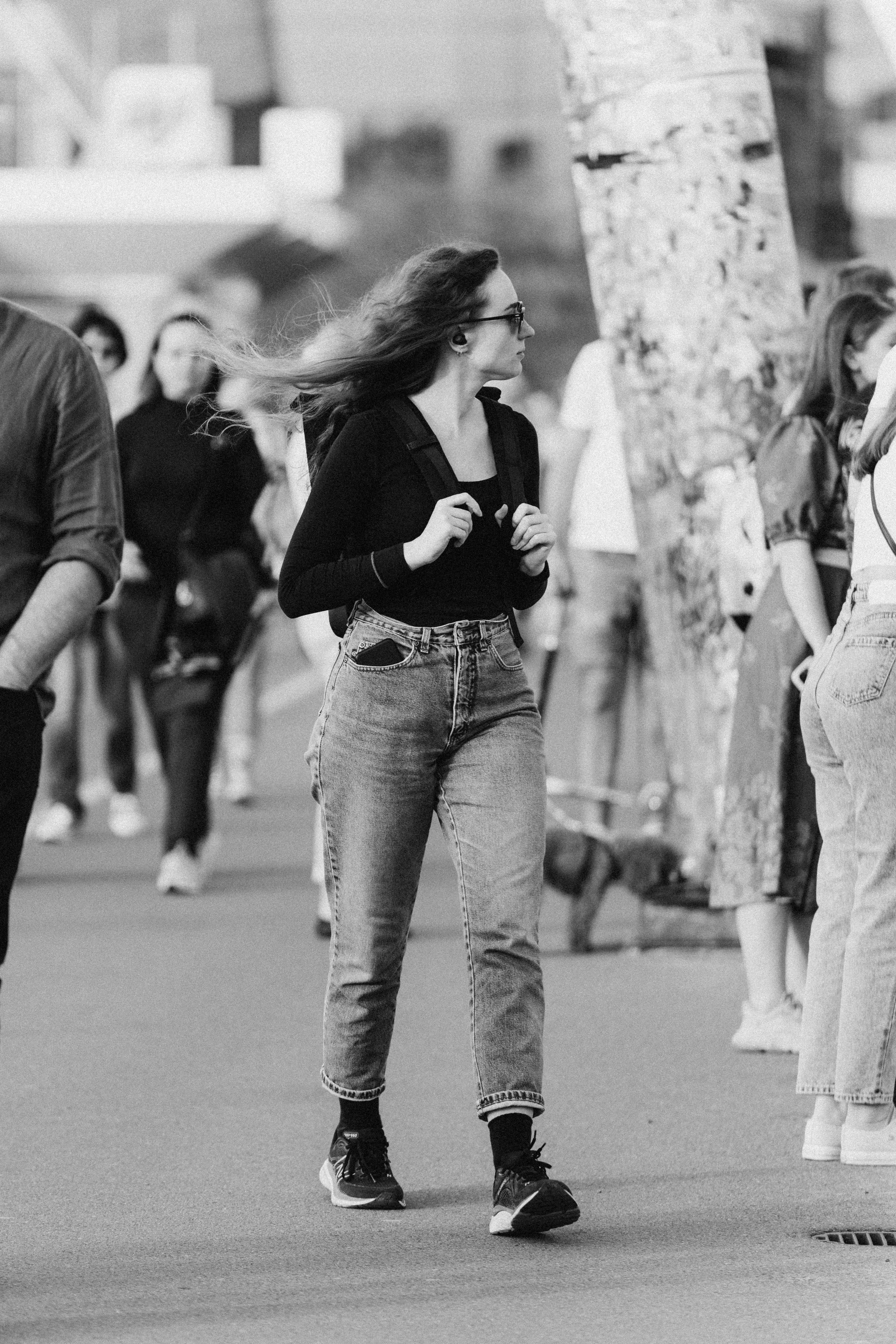 A young woman with long hair and glasses, wearing a black top, jeans, and sneakers, walking outdoors among other pedestrians.