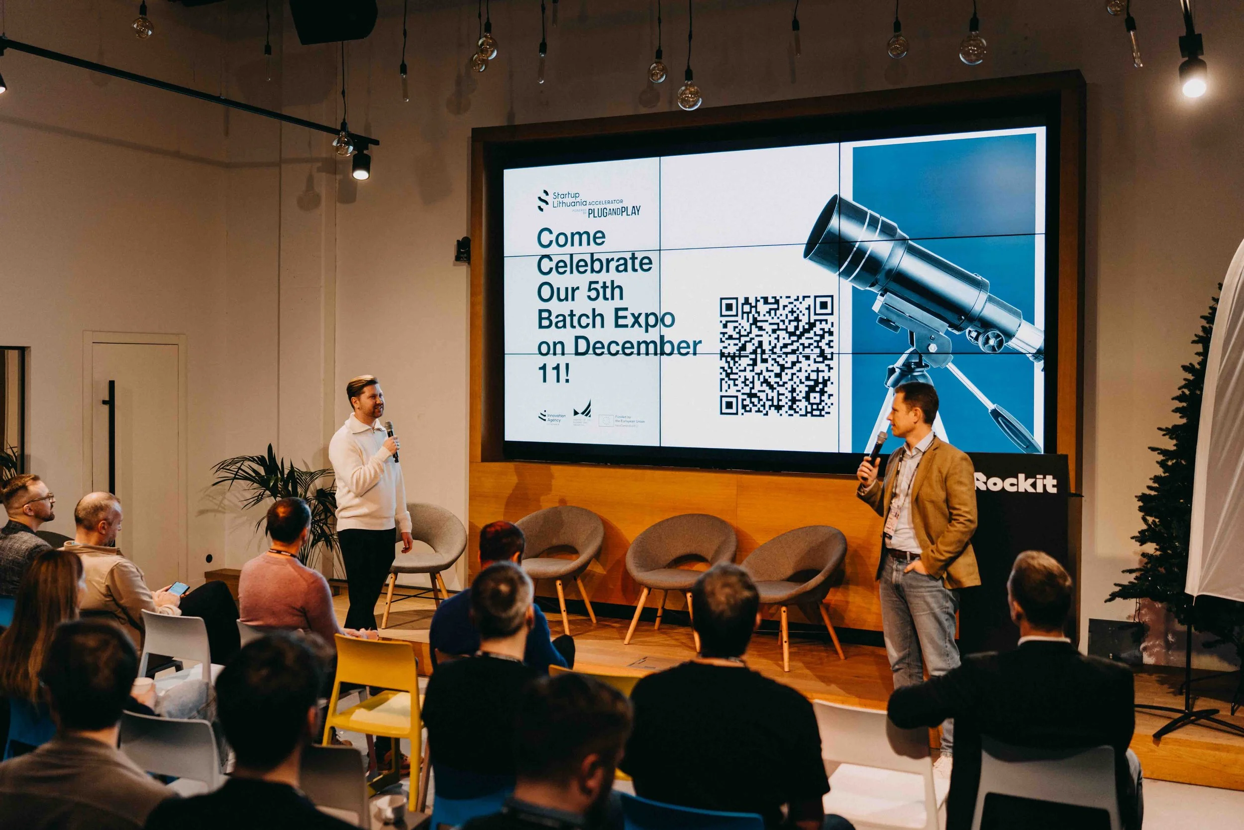 A conference room with an audience listening to two speakers presenting on stage. The large screen behind them displays a promotional message for a 5th Batch Expo happening on December 11, including a QR code. The room has modern decor with plants an