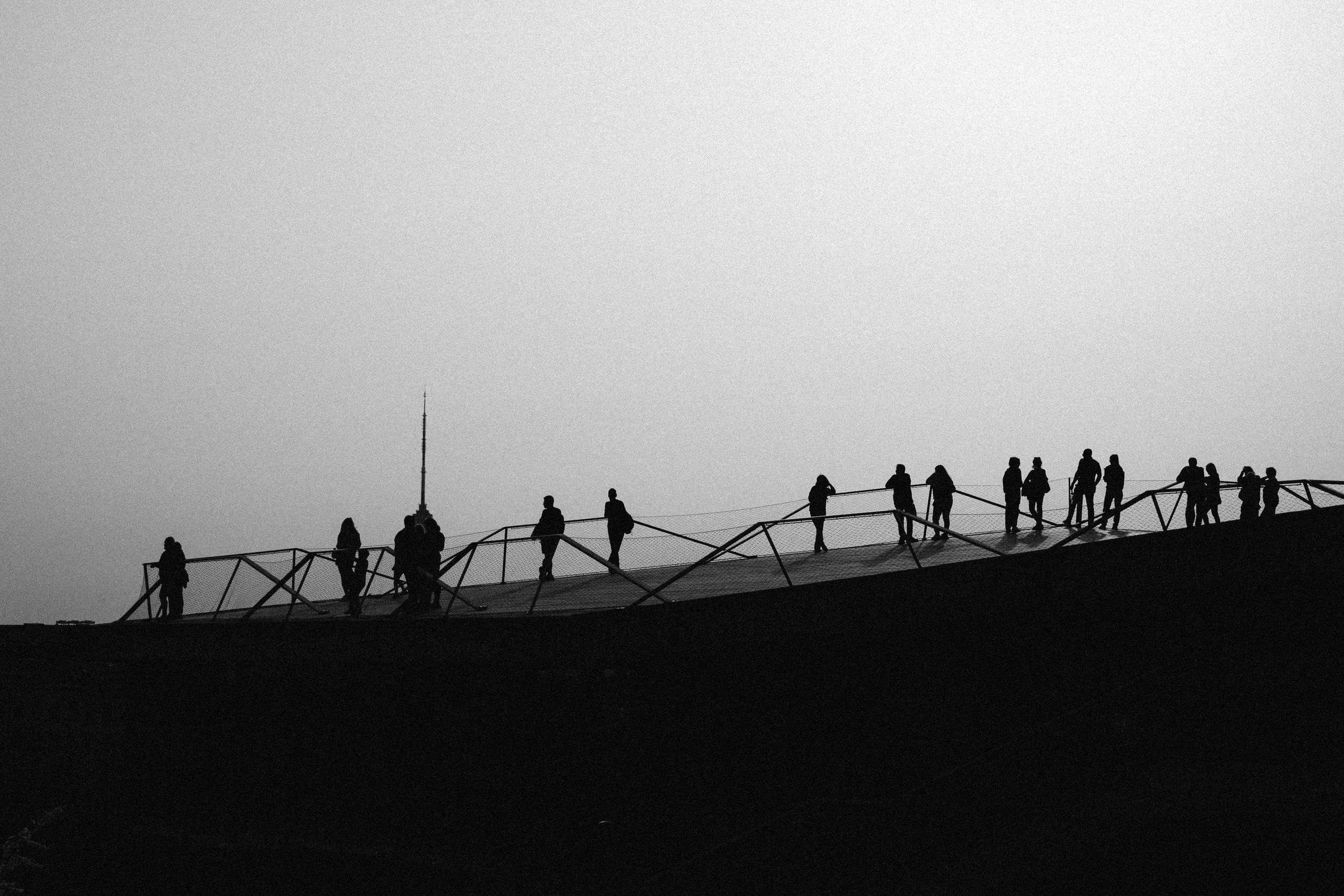 Silhouettes of people walking on a bridge or walkway on a hilltop with a tall antenna or tower in the background, black and white