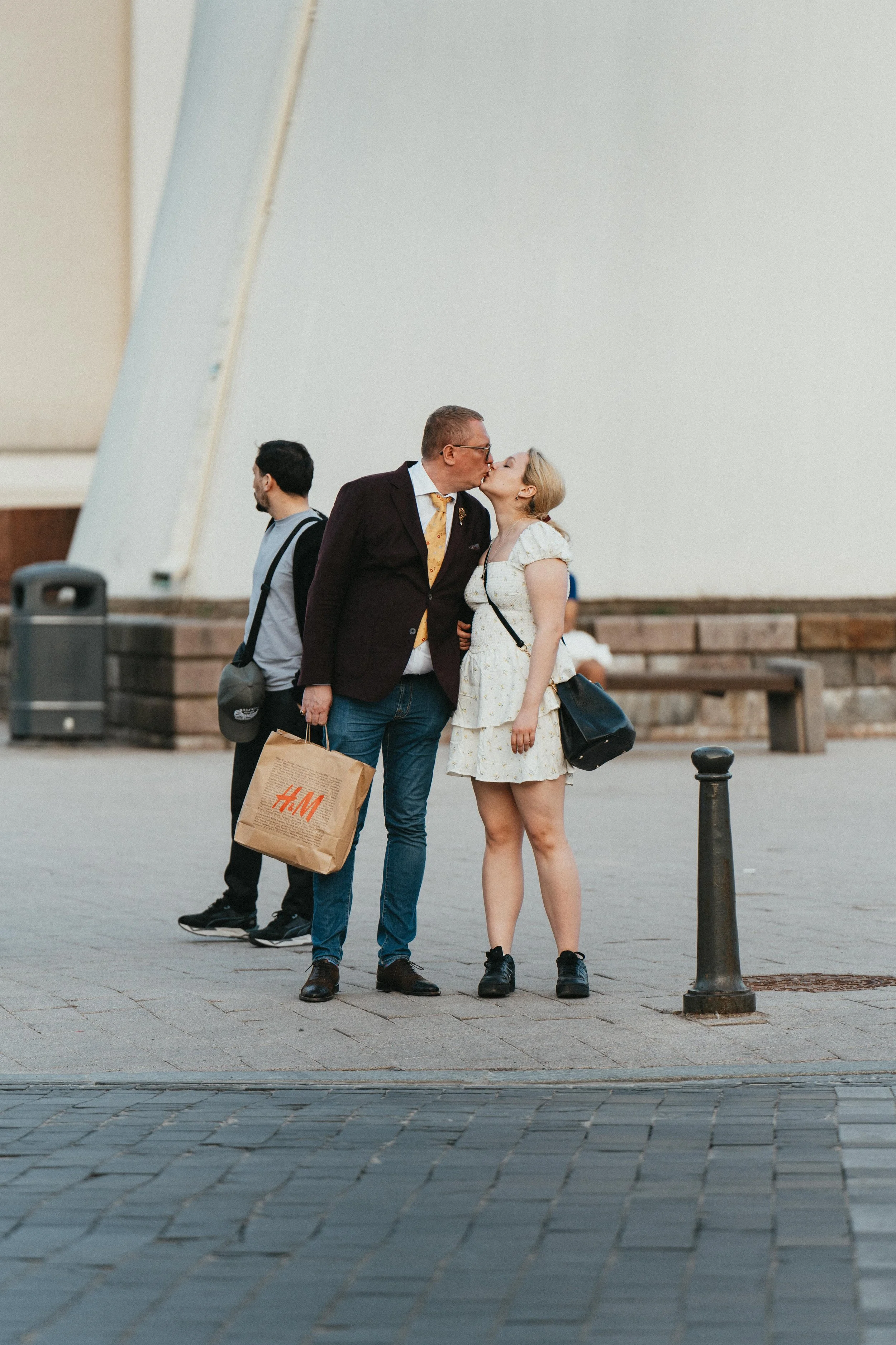 A couple kissing on the street, with a man in the background carrying an H&M shopping bag. The woman wears a white dress, and the man wears a brown blazer and glasses.
