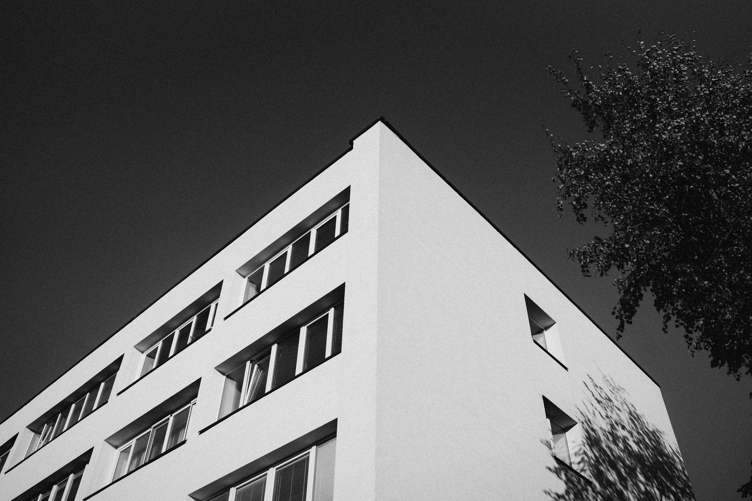 Black and white photo of a modern white building with multiple rectangular windows, taken from a low angle during the daytime, with a shadow of a tree on the side of the building and a dark sky background.