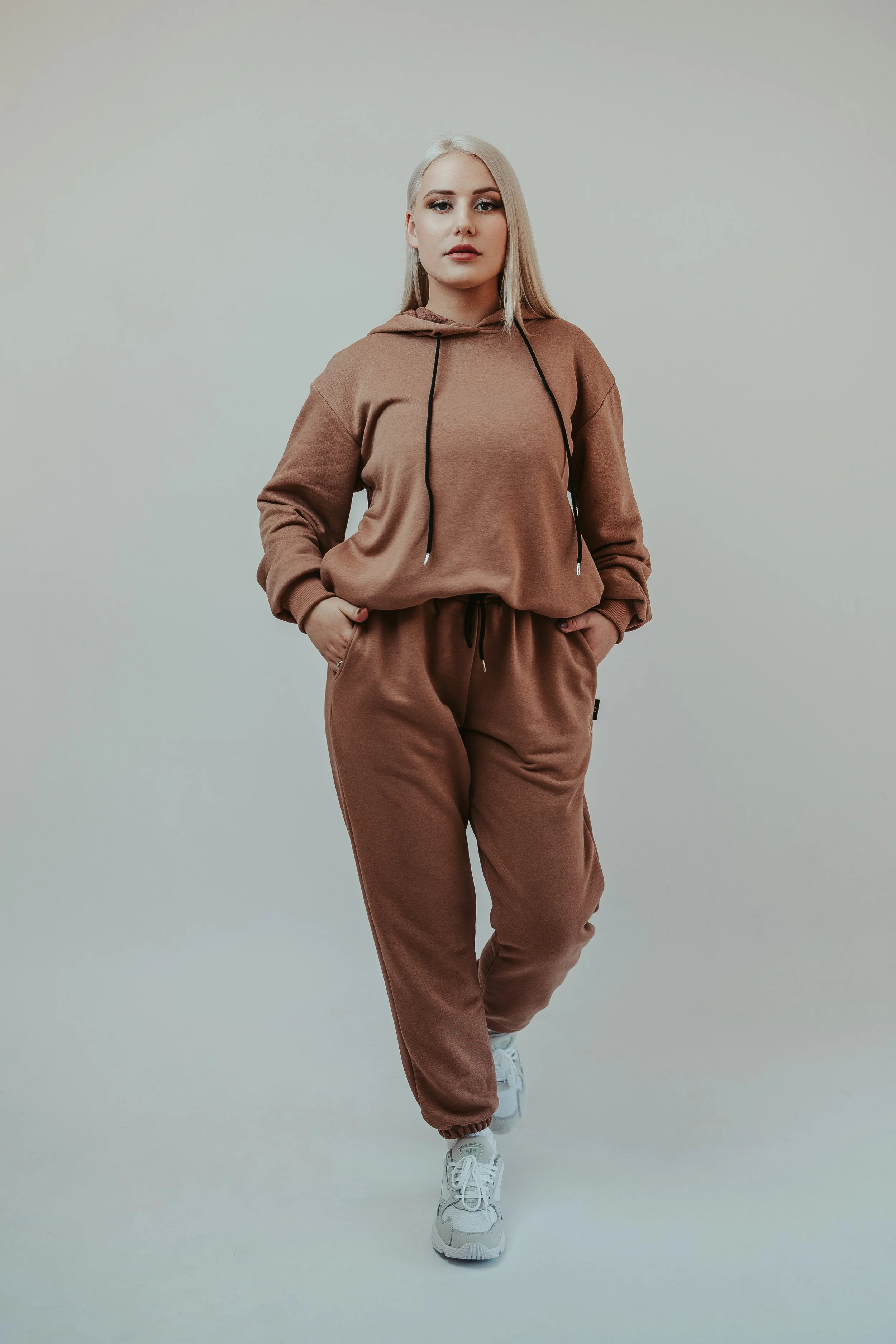 A woman with long blonde hair wearing a brown hoodie and matching sweatpants, standing against a plain light-colored background.