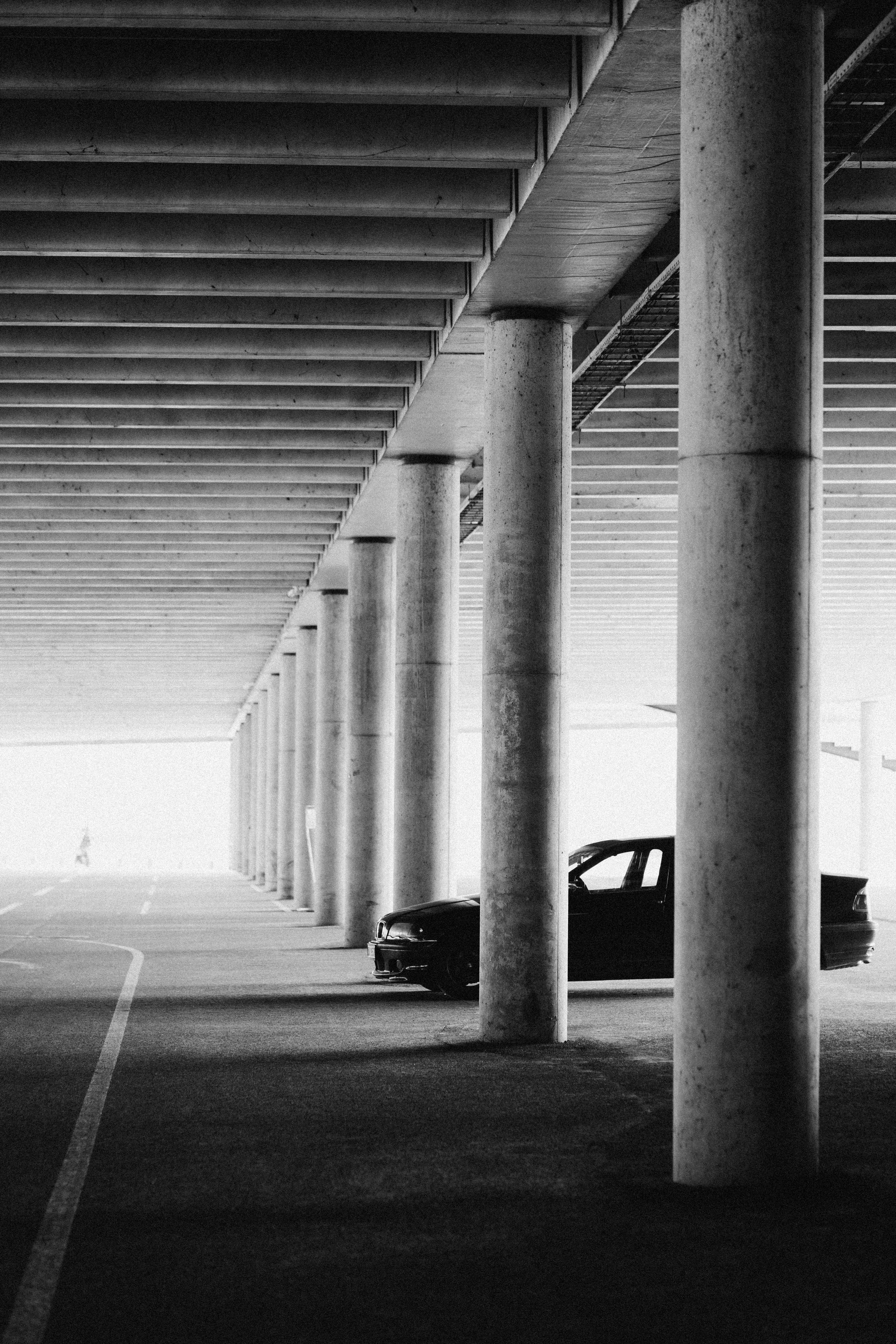 A black car parked under a concrete overpass with tall support columns in black and white.