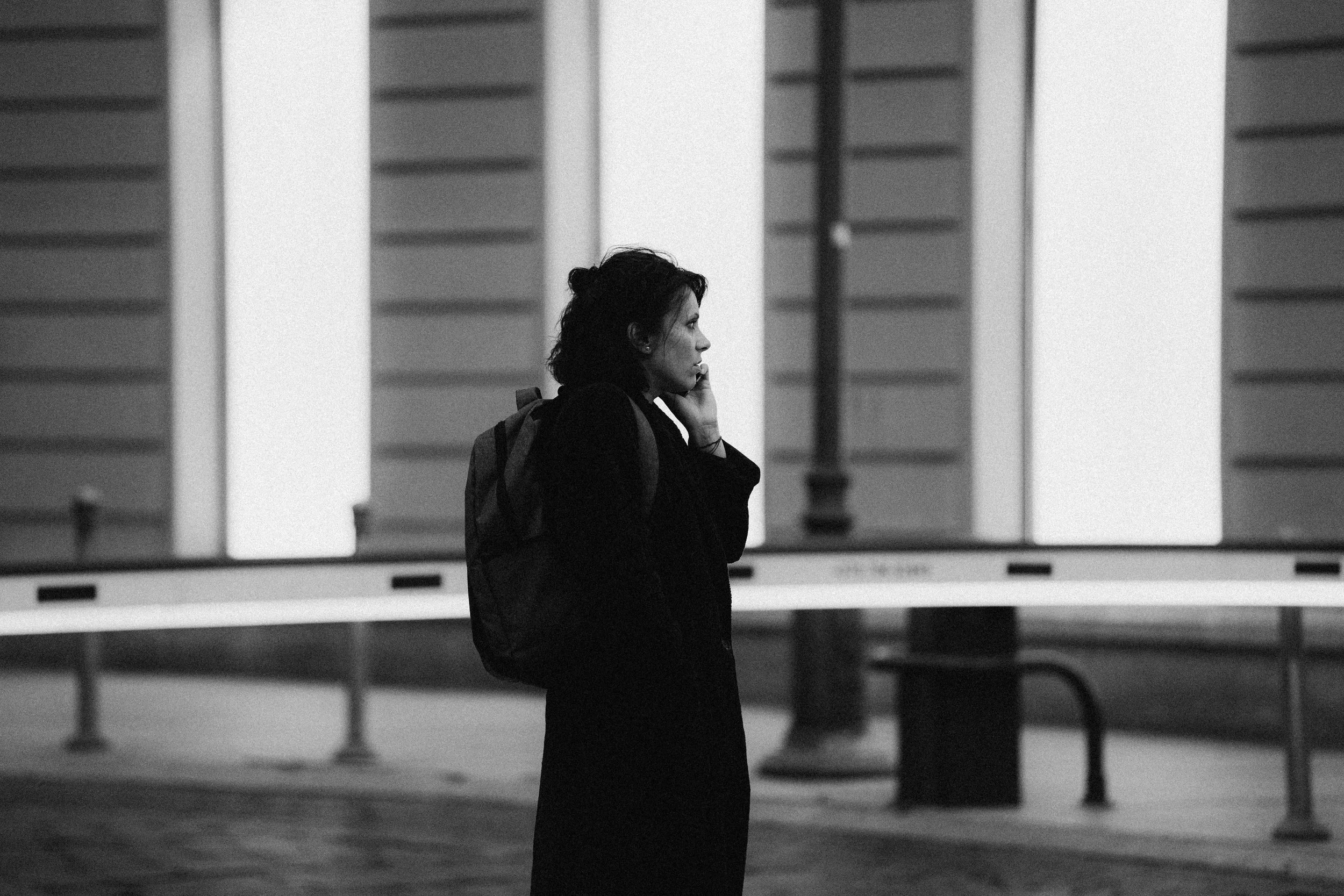 A woman stands on a city street at night, talking on her phone. She has a backpack and is dressed in dark clothing, with buildings and street furniture in the background.