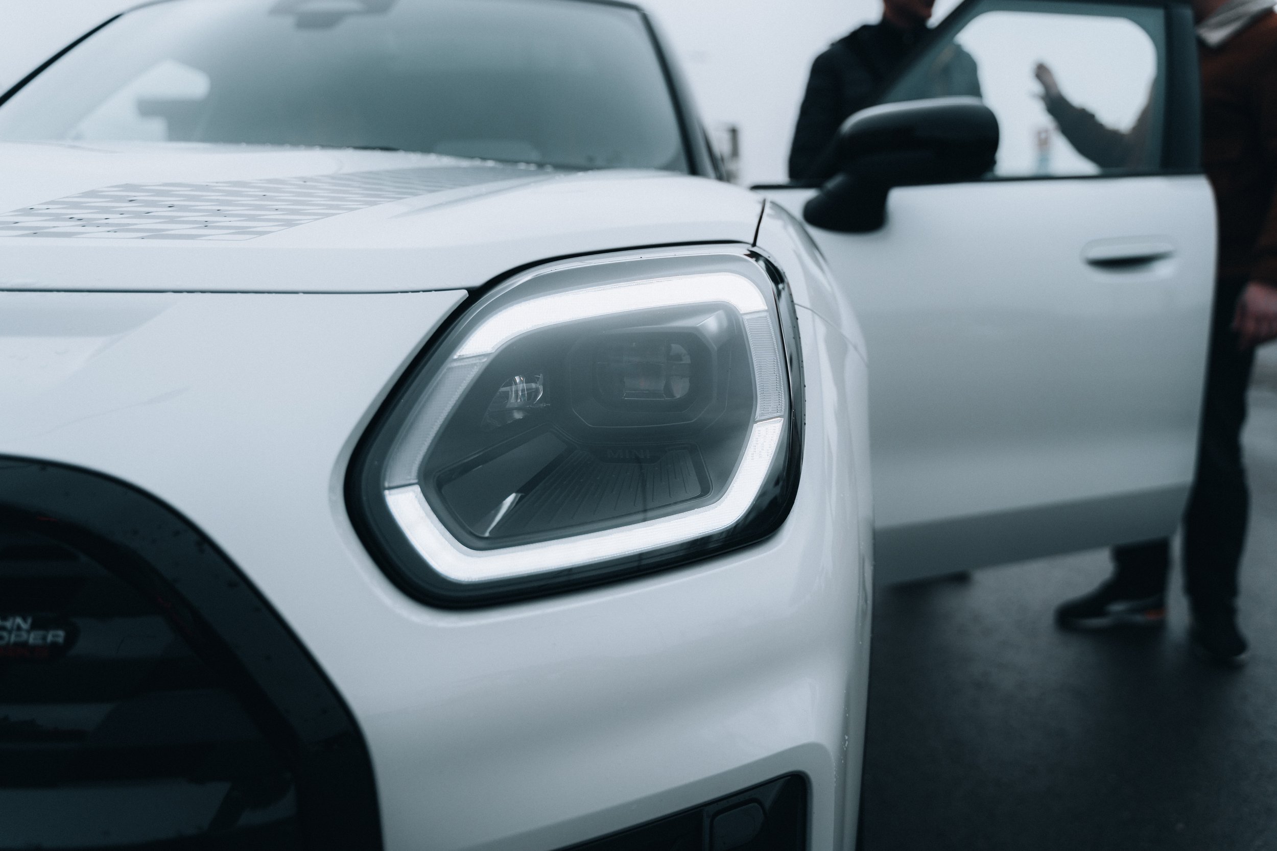 Close-up of a white vehicle's front headlight and open driver-side door, with people standing near the car.