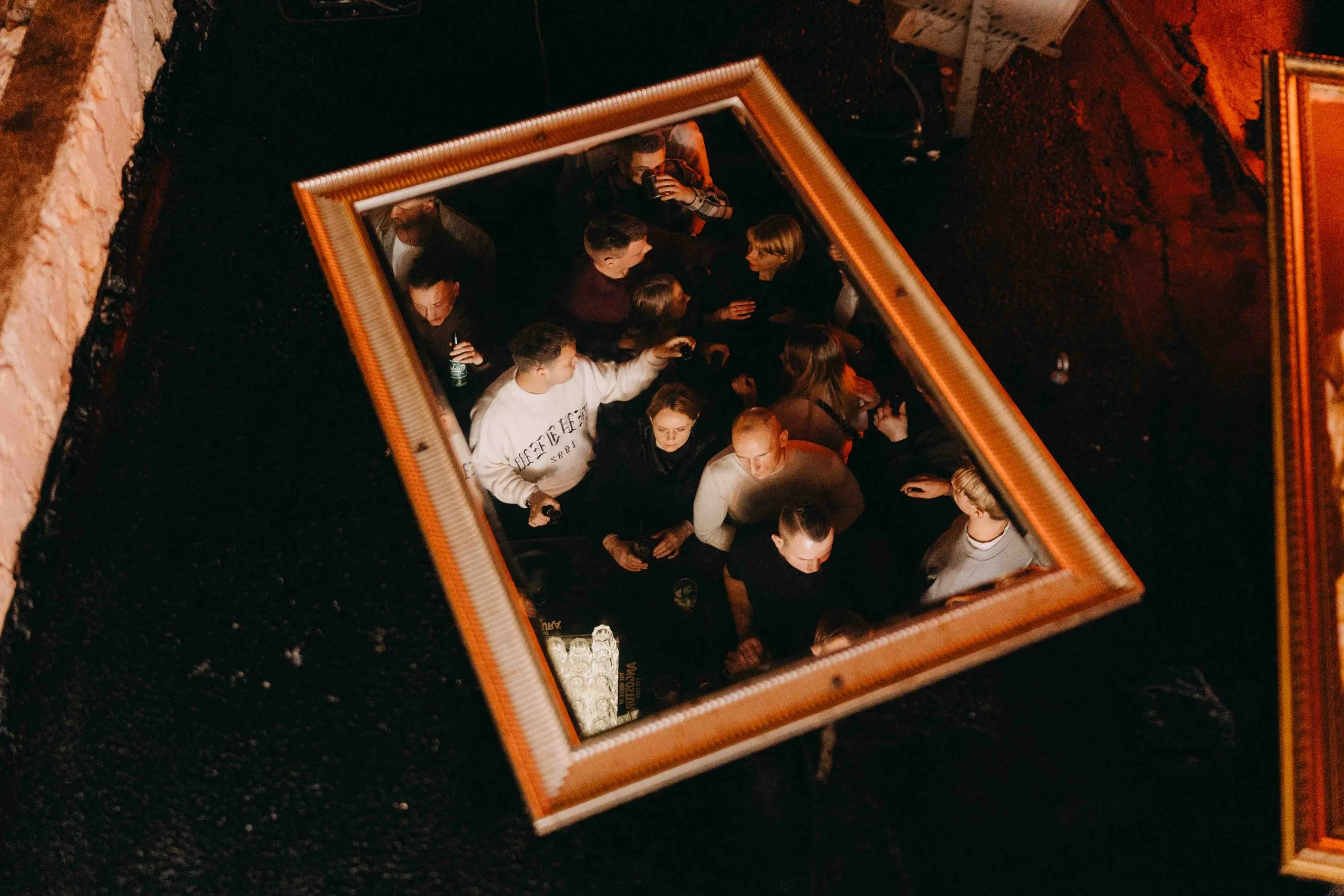 A group of people gathered in a dimly lit room, seen from above through a square mirror on the ceiling.