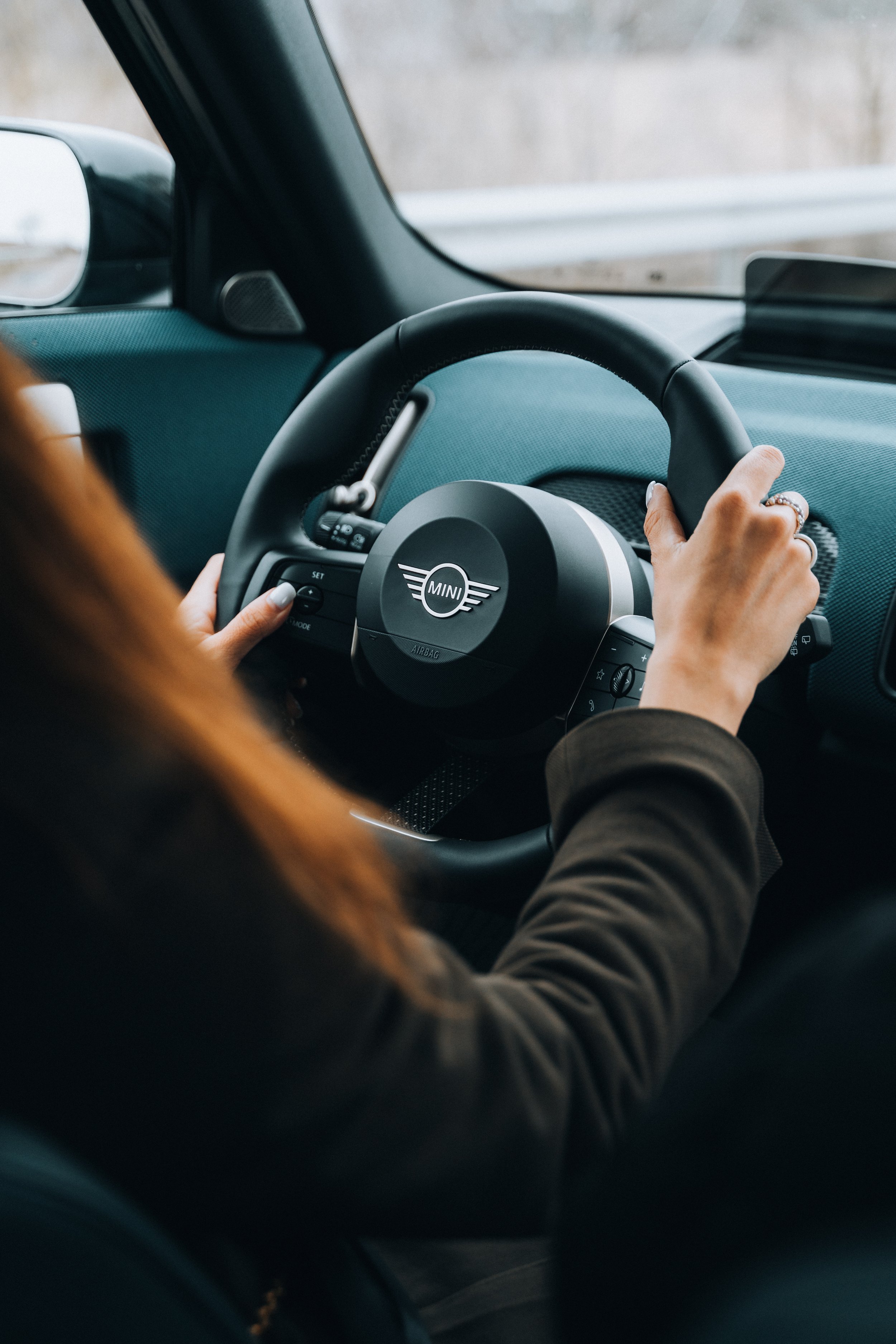 A person driving a car with a Mini Cooper logo on the steering wheel, seen from the perspective of the passenger seat.