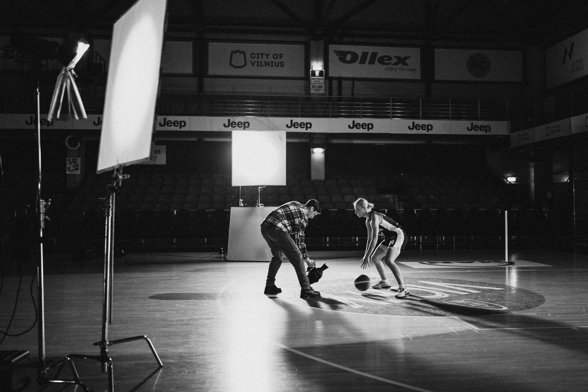 A woman and a man practicing basketball on an indoor court with studio lights and equipment set up around them, empty stadium seating in the background.