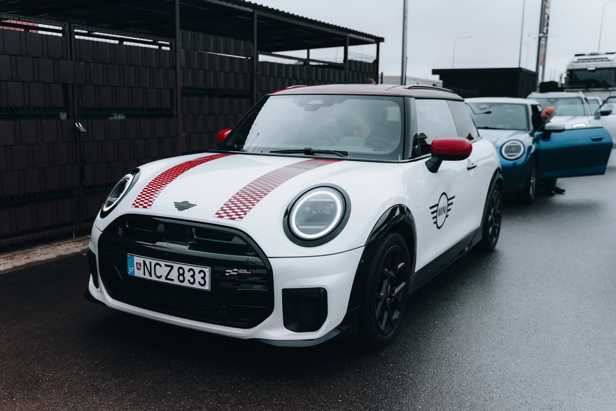 A white Mini Cooper with red and gray checkered racing stripes and red side mirrors, parked on a wet road, with other cars in the background.