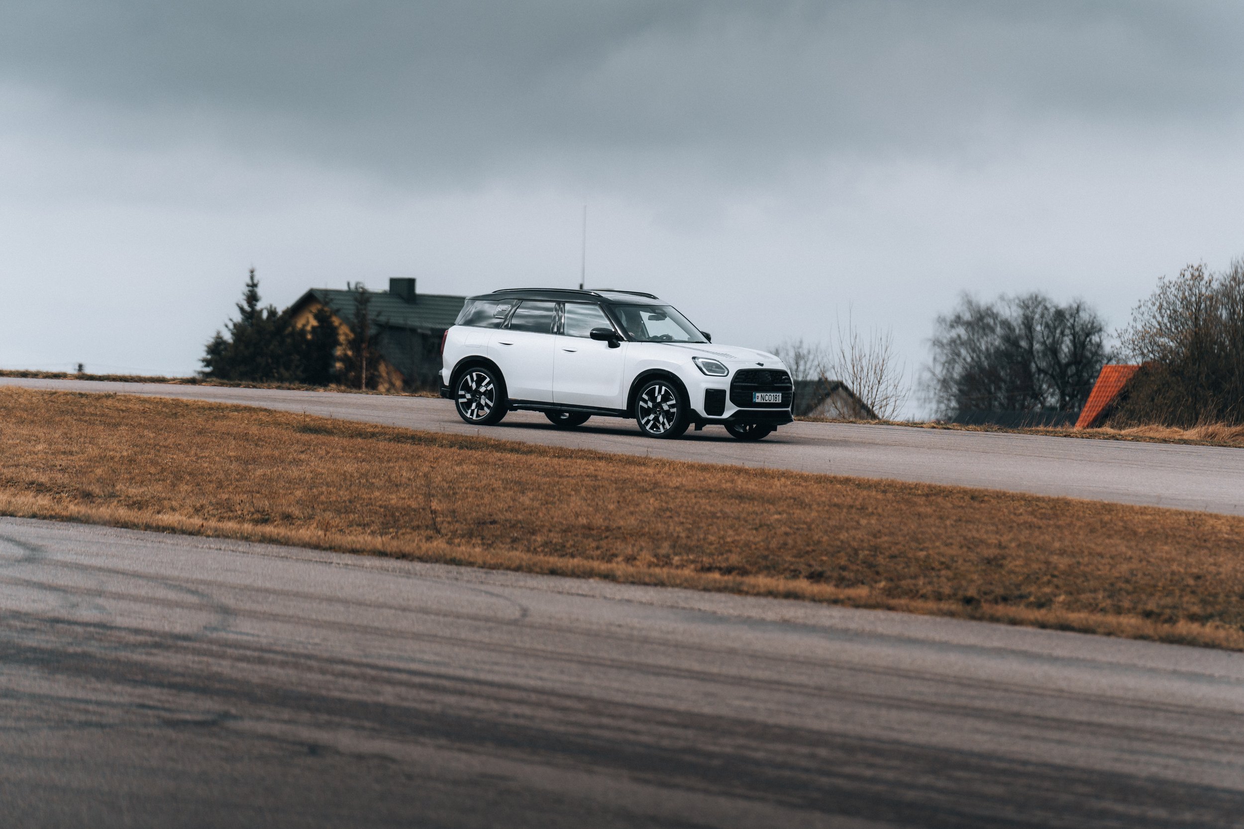 A white SUV driving on a paved road in a rural area with houses and trees under a cloudy sky.
