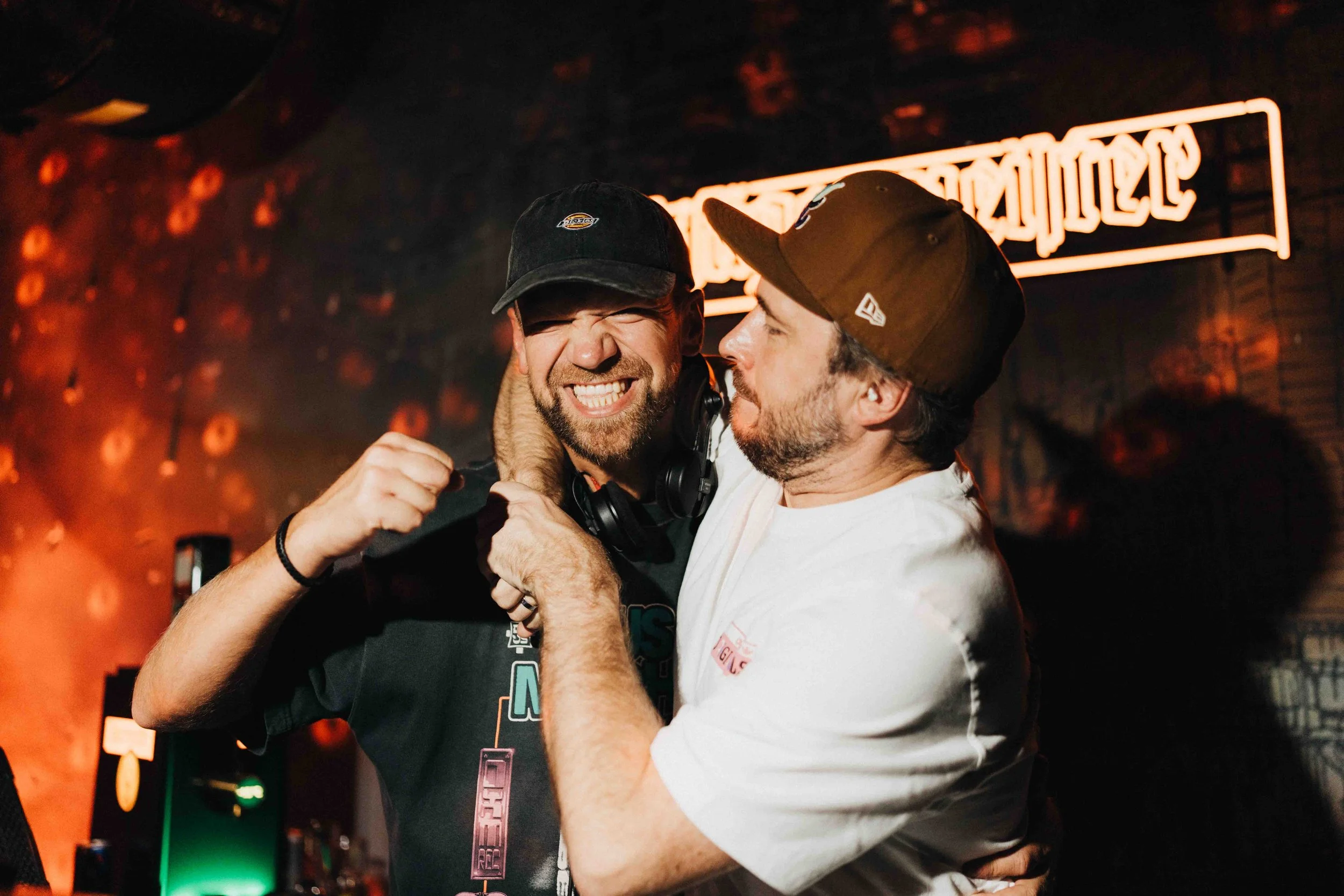 Two men celebrating and smiling together in a lively indoor setting with neon lights in the background.