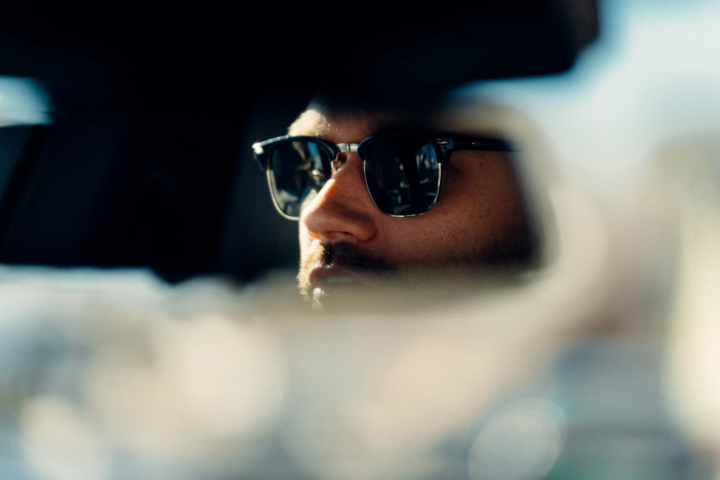 A man with dark hair and sunglasses looking out from a rearview mirror of a car.