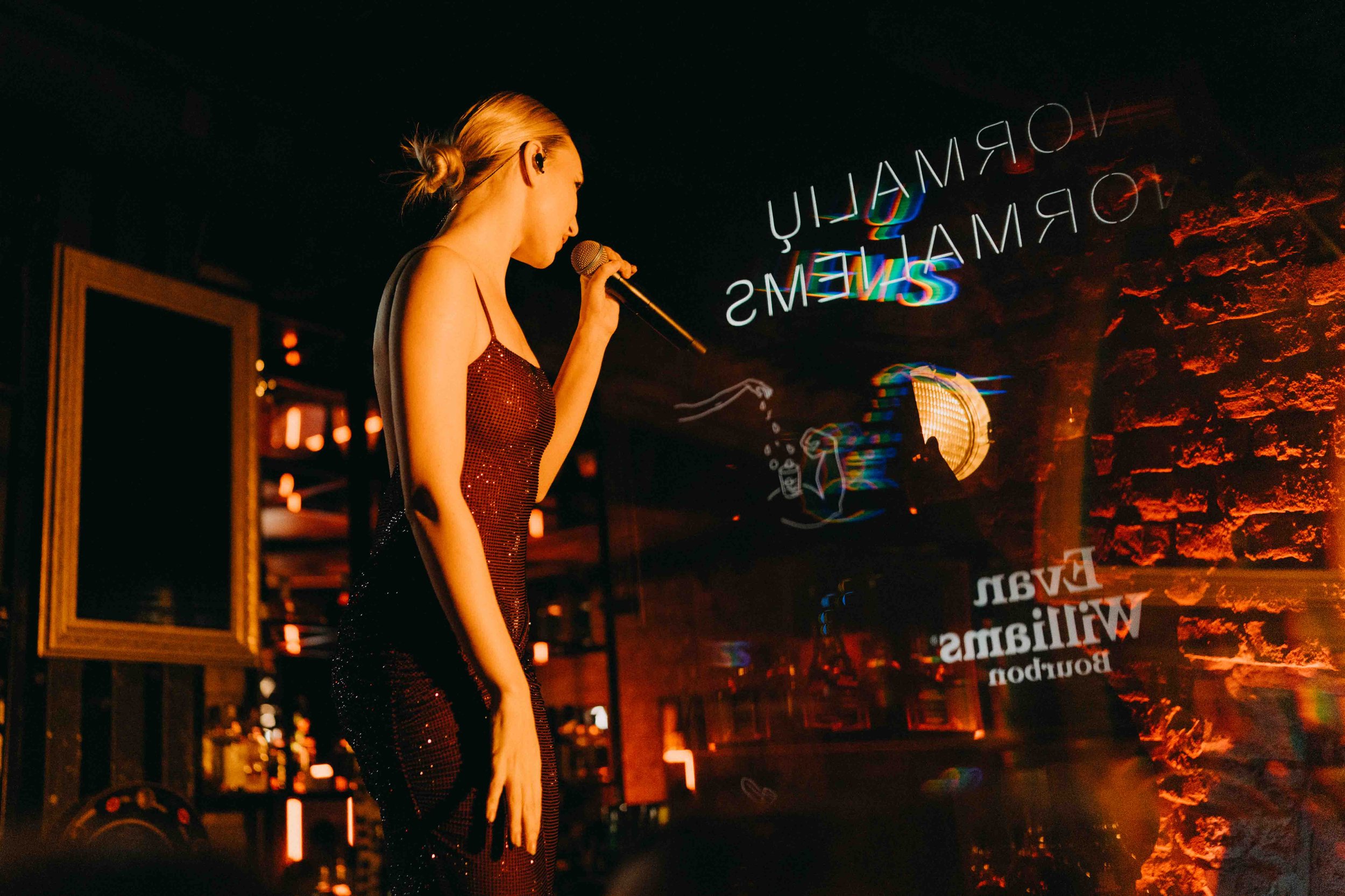 A woman with blonde hair in a ponytail sings into a microphone at a bar or nightclub. She is wearing a sleeveless, red, sequined dress. The background features a brick wall and neon signs displaying the words 'VOLUME', 'ROMAM', and 'WILLIAMS'. There 