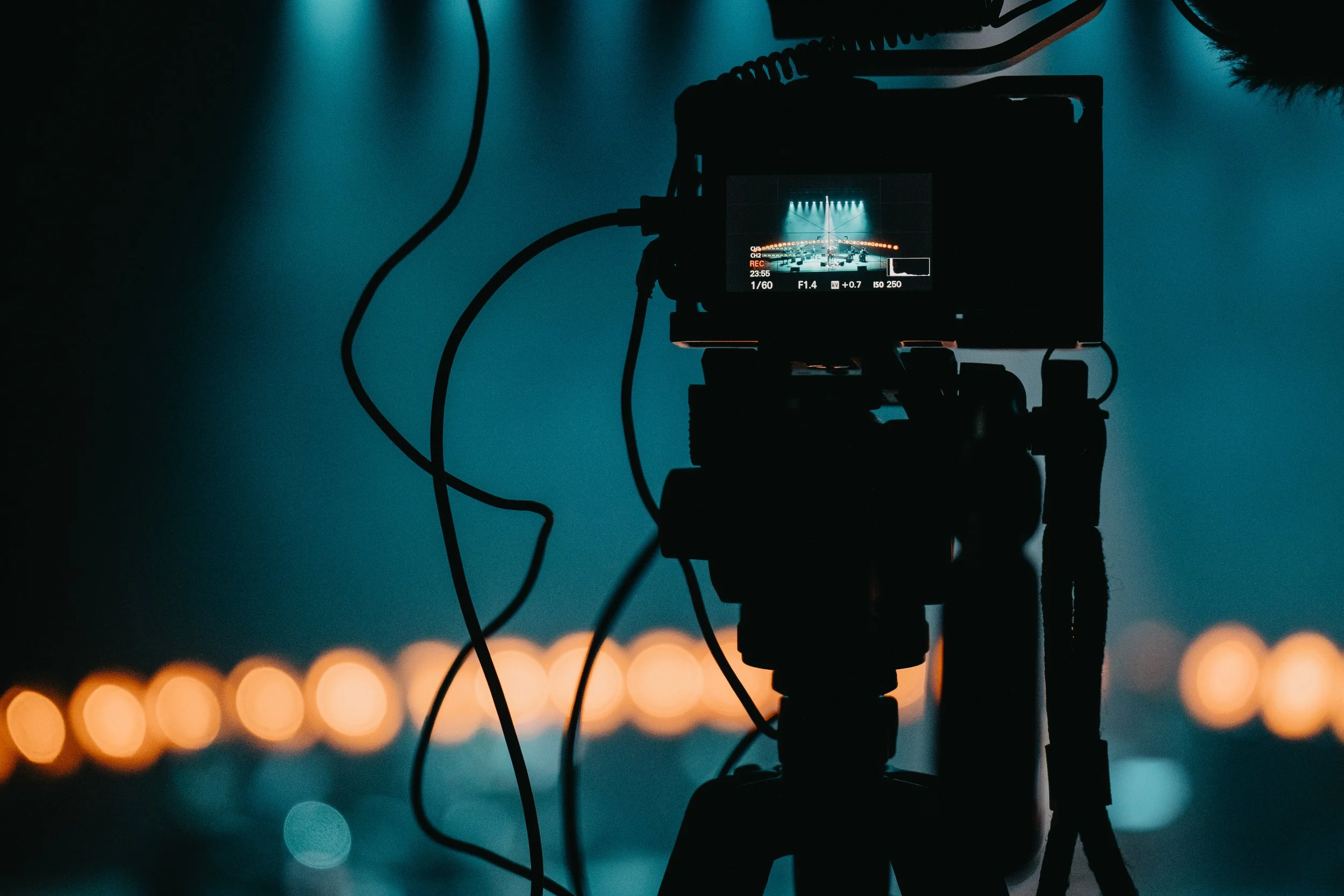 A camera mounted on a tripod captures a concert stage with spotlights and hanging lights in the background.