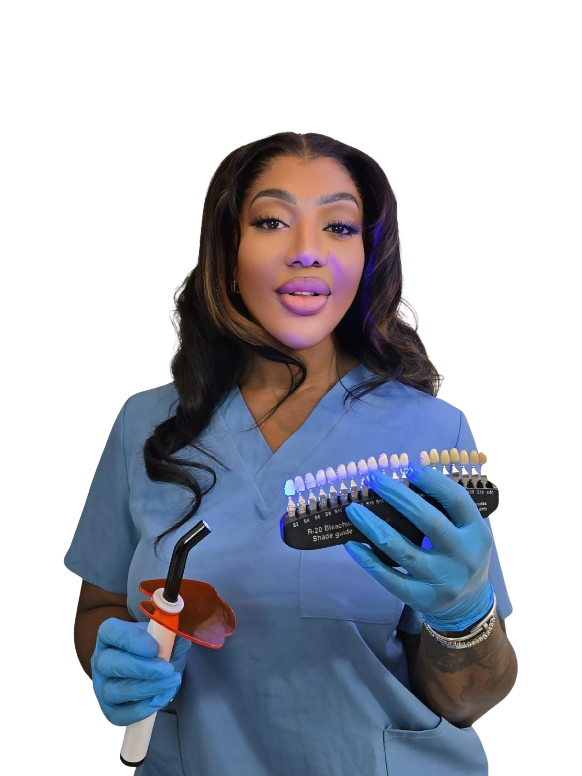 A woman wearing blue dental scrubs and gloves, holding dental shade guides in one hand and a dental curing light in the other, standing against a plain background.