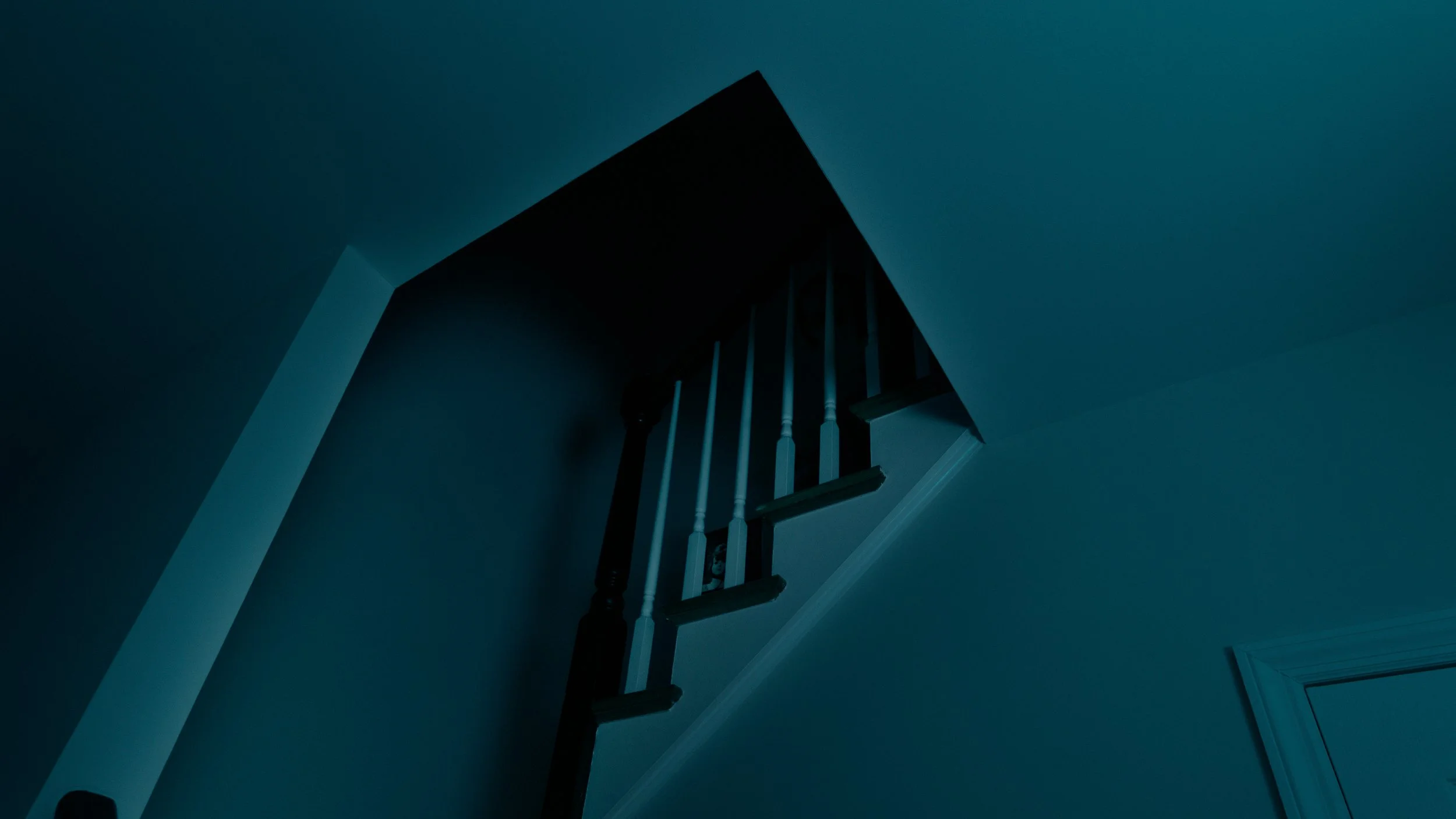 Dark staircase leading up to an attic or upper floor, with wooden railing and support beams, in dim lighting, with something hidden in the shadows.
