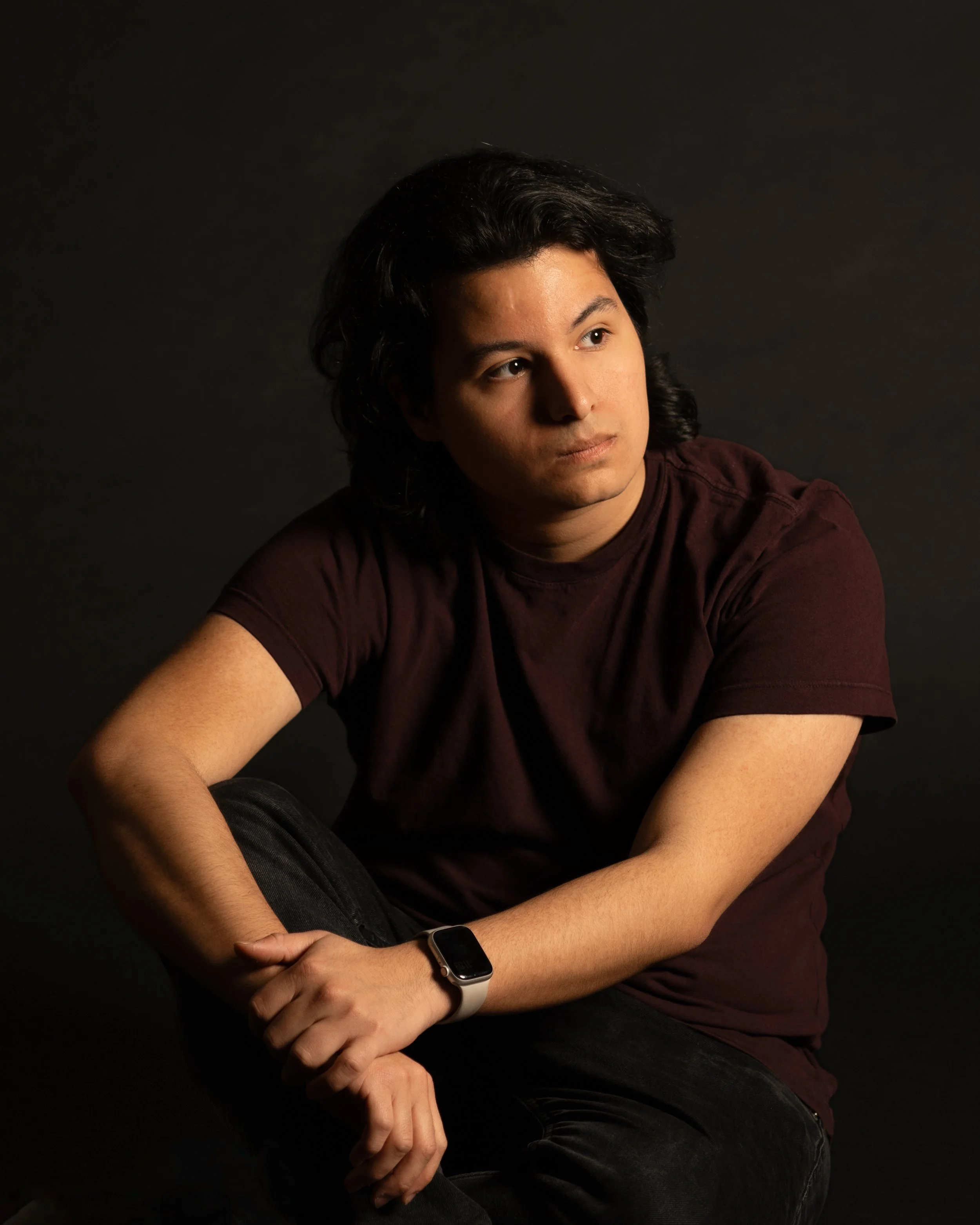 A young man with dark hair, wearing a dark maroon T-shirt and a smartwatch, sits against a black background, looking to the side.