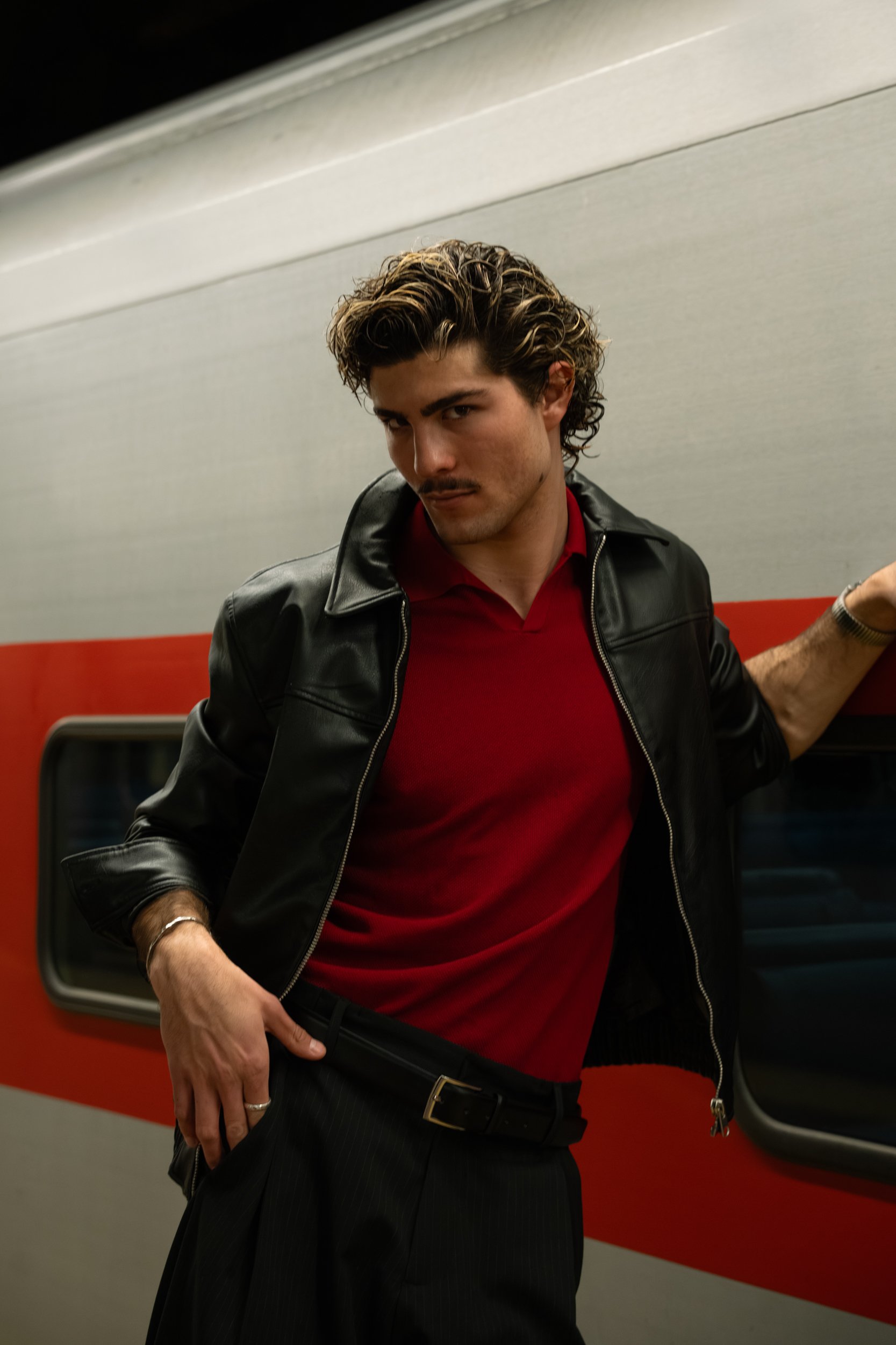 A young man with styled hair, a mustache, and light skin, posing on a train, wearing a black leather jacket over a red shirt, with dark striped pants, and sunglasses on his head.