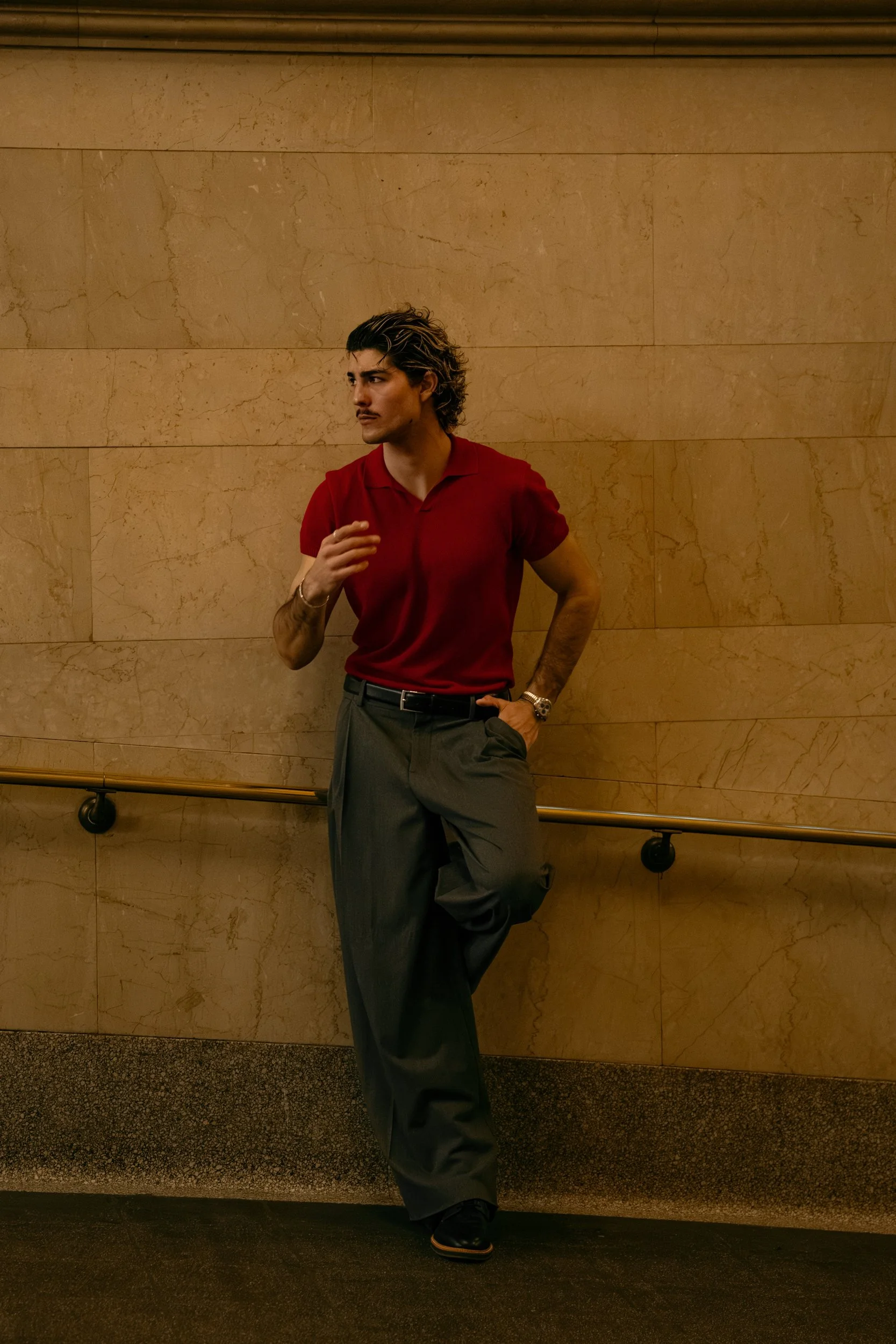 A young man with dark, wavy hair, dressed in a red polo shirt and gray pleated trousers, leans against a beige marble wall with his left hand in his pocket while his right hand is slightly raised. He is standing near a horizontal handrail in an indoo