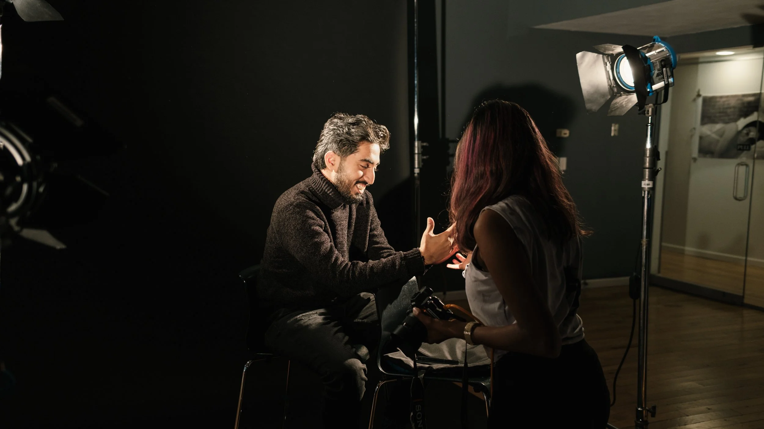 A man and a woman interact on a film or photo set; the man is sitting on a chair smiling, and the woman is standing, possibly acting or directing. Studio lighting is visible.