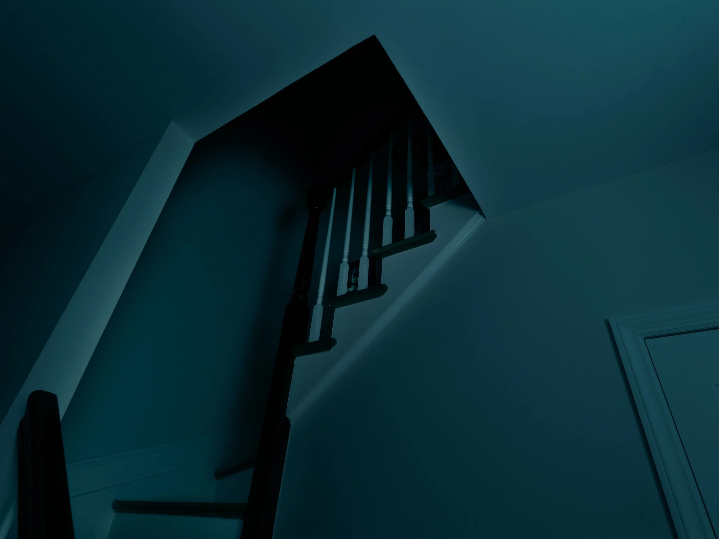 Dark staircase leading up to an attic or upper floor, with wooden railing and support beams, in dim lighting, with something hidden in the shadows.