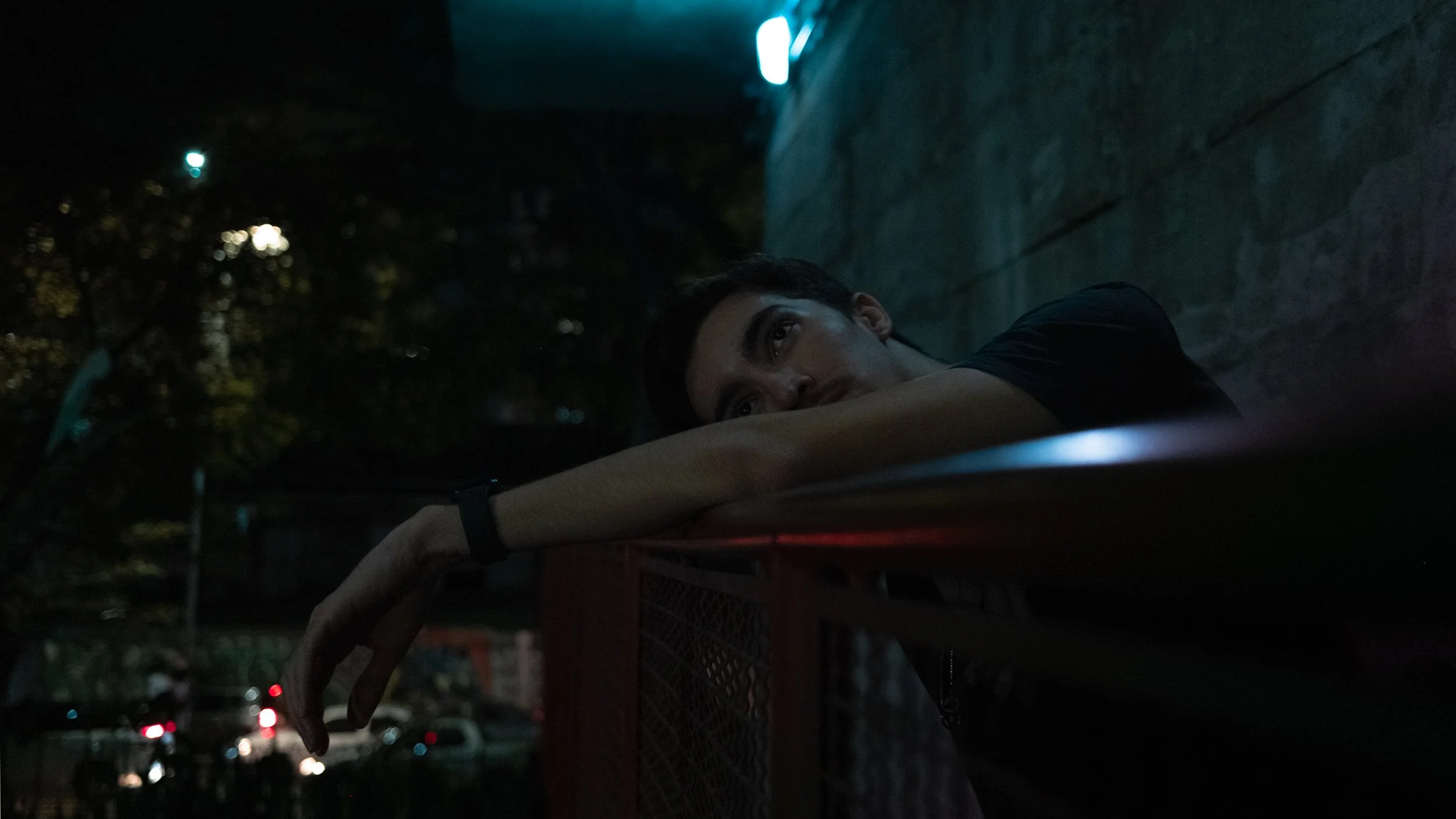 Young man resting his arm on a red metal fence at night, gazing upward with a contemplative expression, in a dimly lit outdoor setting.