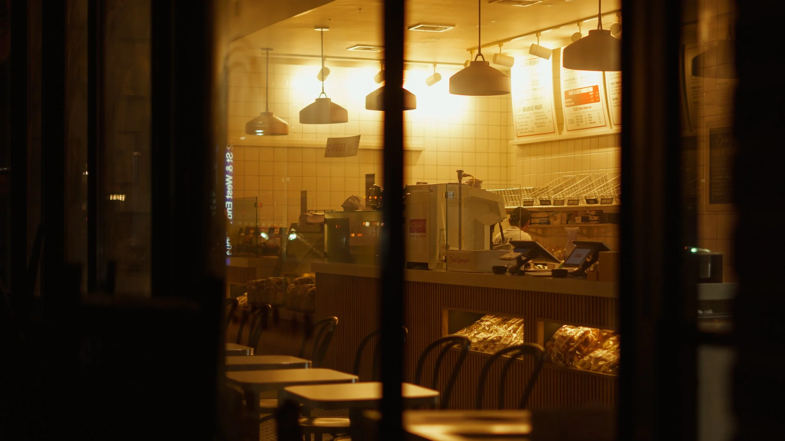 Scene of a café or bakery viewed through a window at night, showing chairs and a warm-lit counter with menu boards overhead.