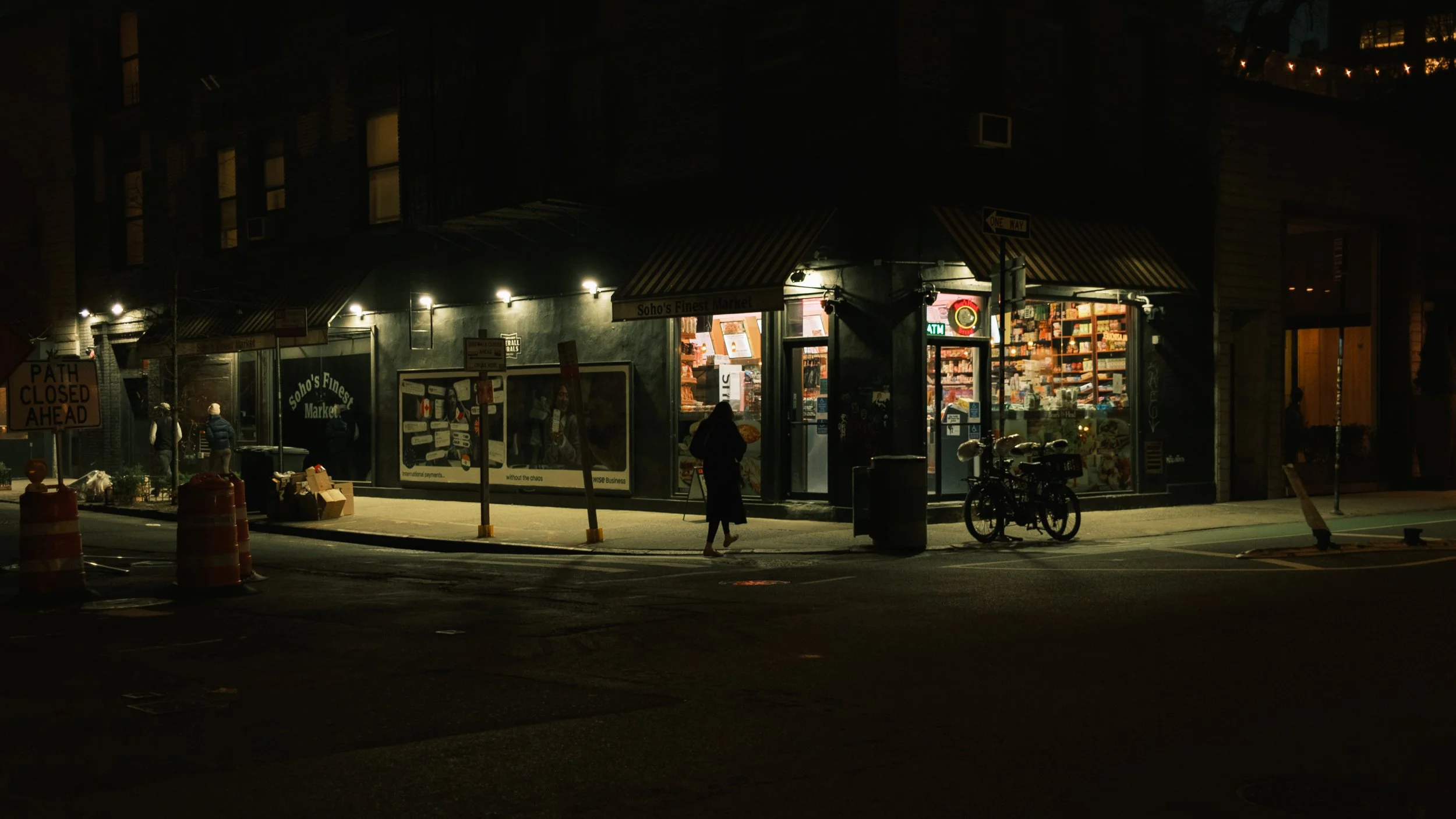 Nighttime scene of a small grocery store named 'Soho's Finest Market' with lights on, a person walking towards the entrance, bicyclists parked outside, and traffic cones on the street.