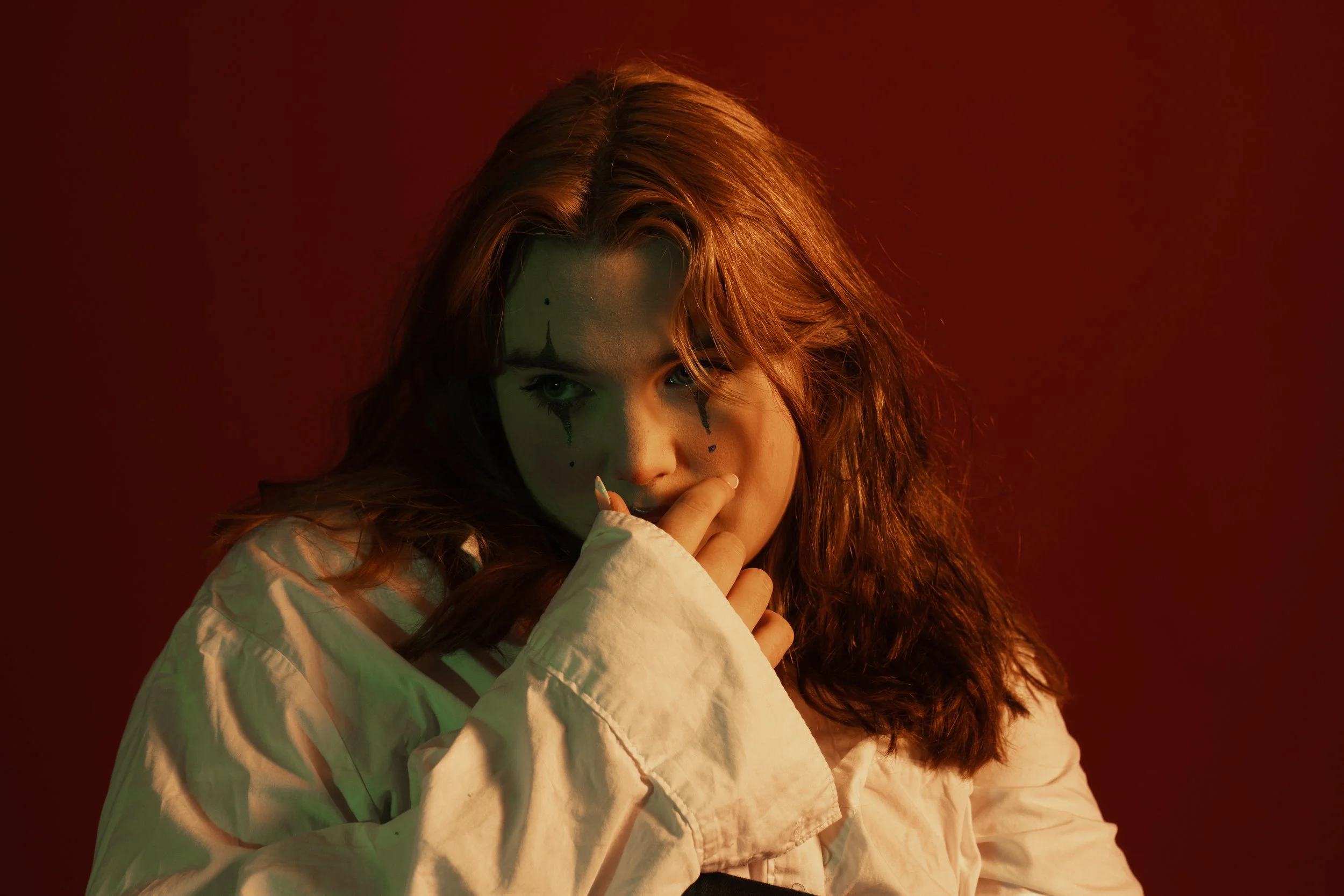 Young woman with long red hair and clown face makeup, wearing a white oversized shirt, biting her finger, in a dimly lit setting with red and green lighting.