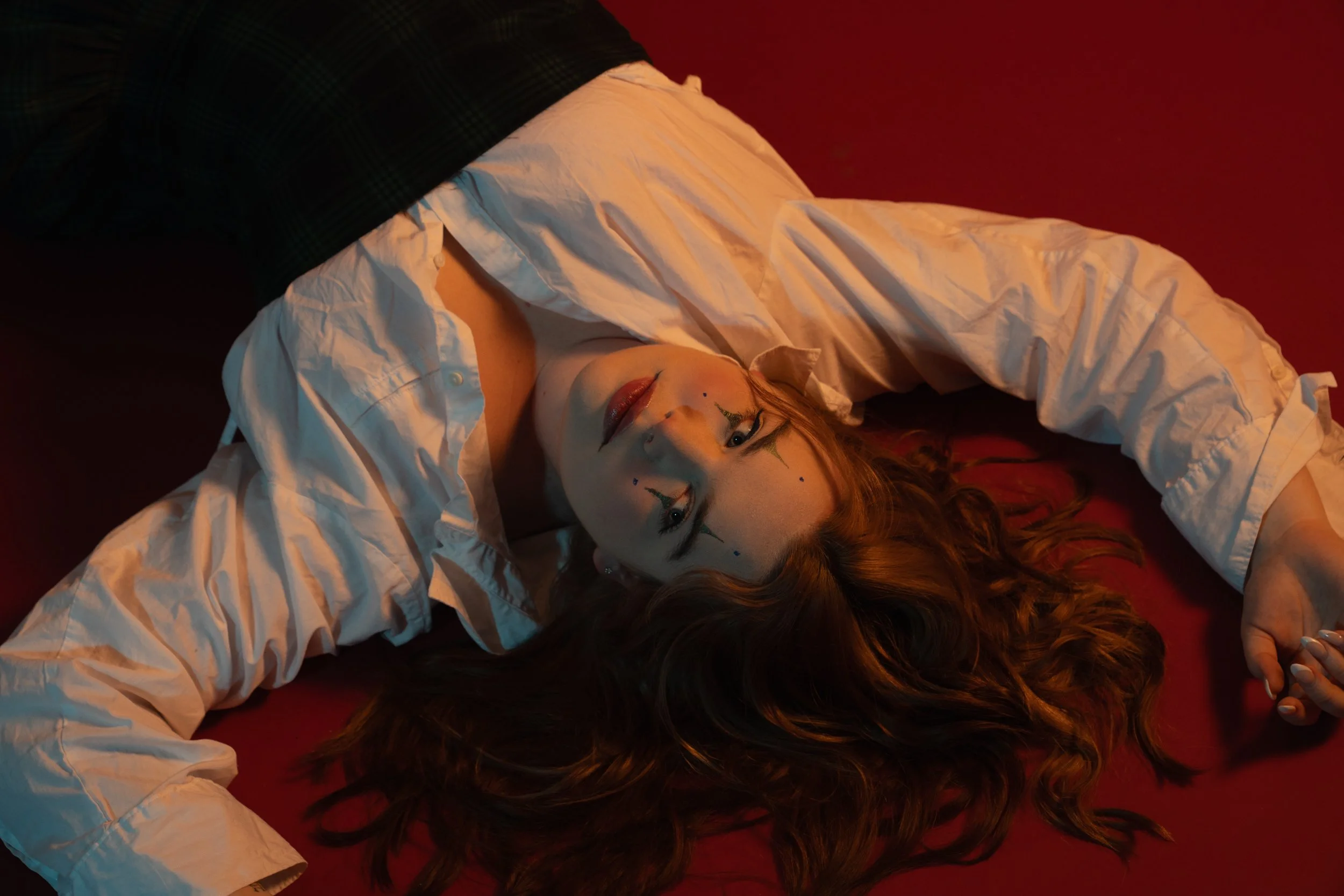 A woman with long, wavy red hair laying on a red surface, wearing a white button-up shirt, dark pants, and a clown makeup with small decorative markings on her face.