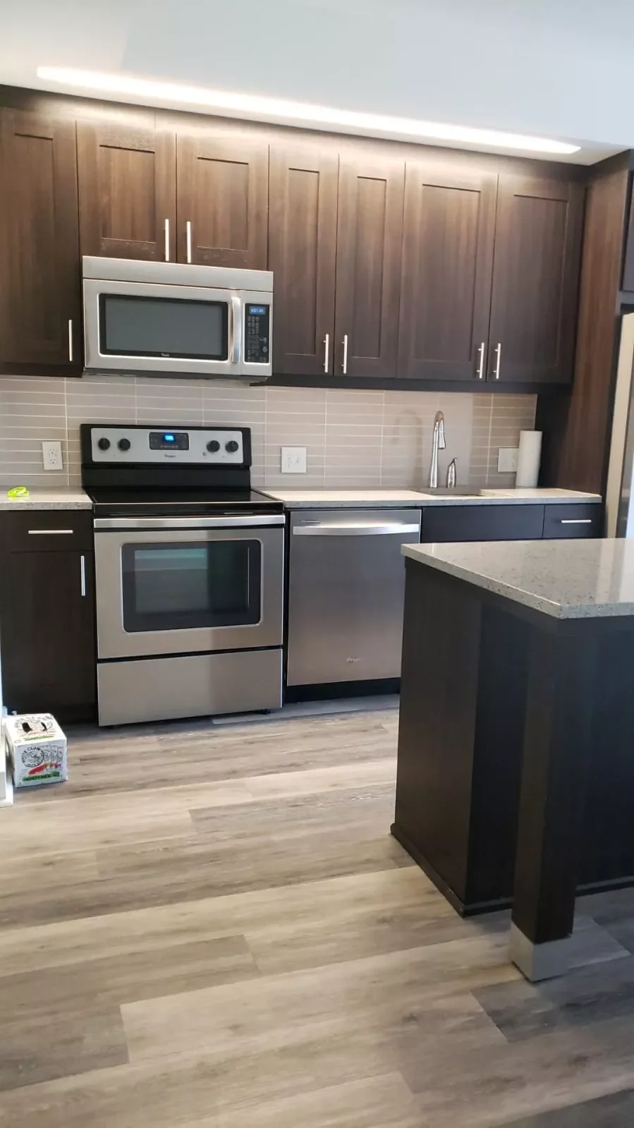 Modern kitchen with dark wood cabinets, stainless steel appliances including microwave, oven, and dishwasher, beige tile backsplash, and a gray speckled countertop.