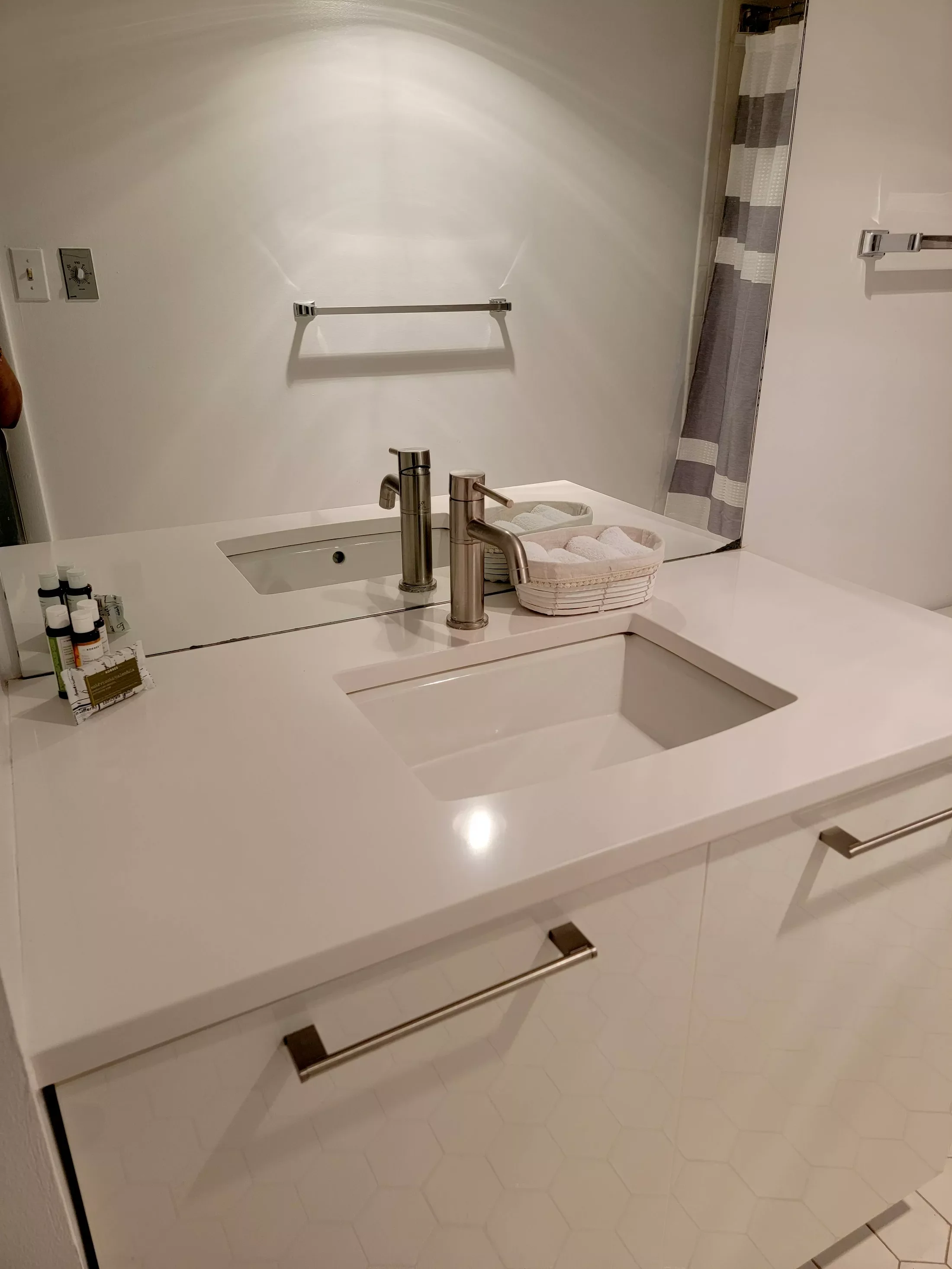 Modern bathroom sink area with a white countertop, a built-in rectangular sink, and a large mirror. There are two brushed nickel faucets, a basket of rolled towels, and toiletry bottles on the counter. A towel bar and a small high shelf are mounted o