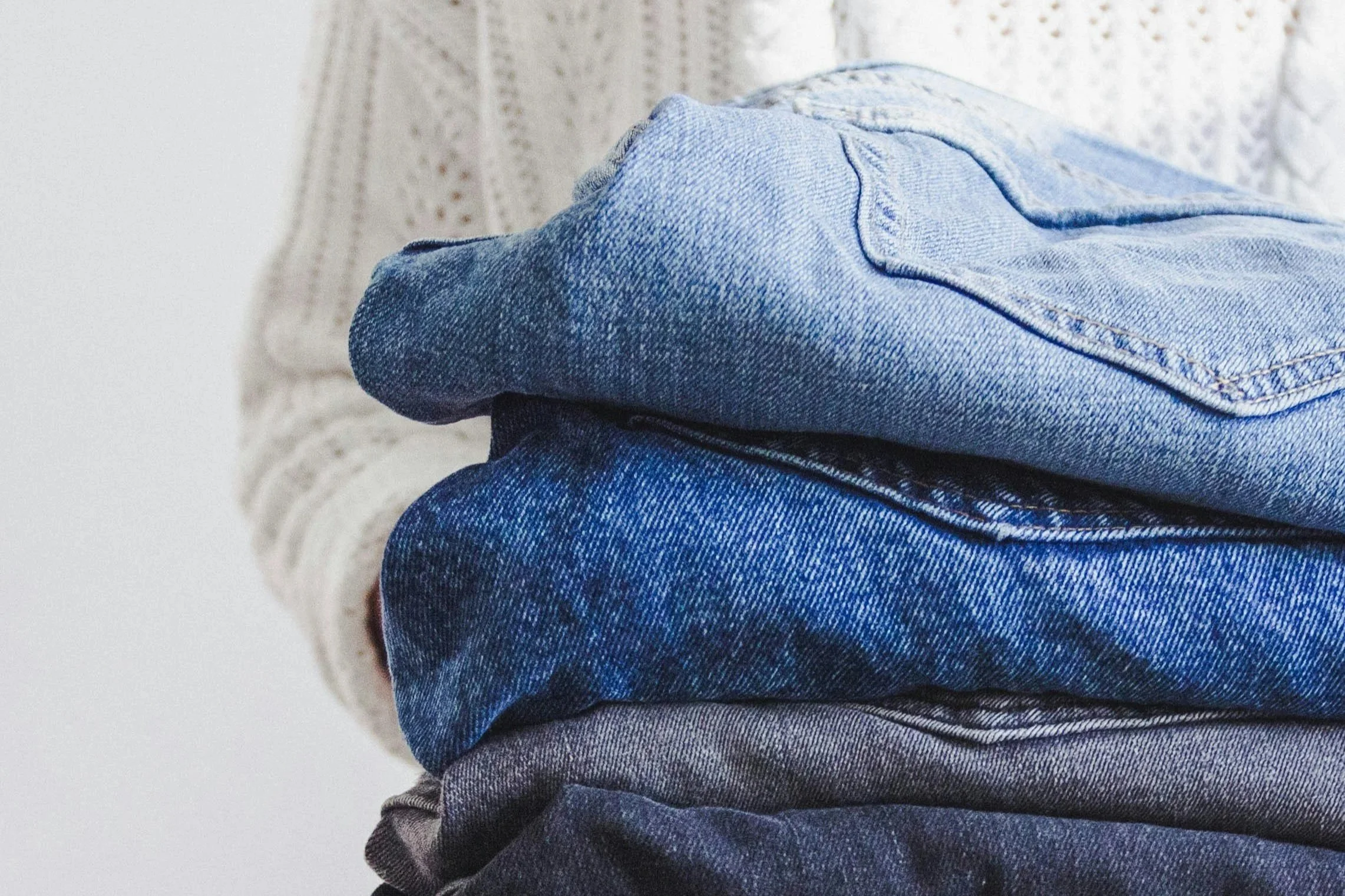 A stack of folded blue and gray denim jeans in front of a person's torso, wearing a knitted cream-colored sweater.