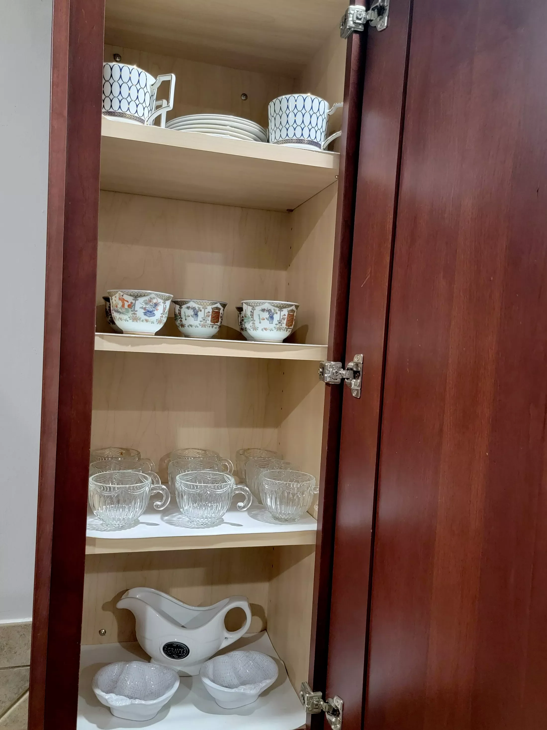 A wooden kitchen cabinet open to reveal shelves with various dishes including mugs, bowls, teacups, and a creamer pitcher.