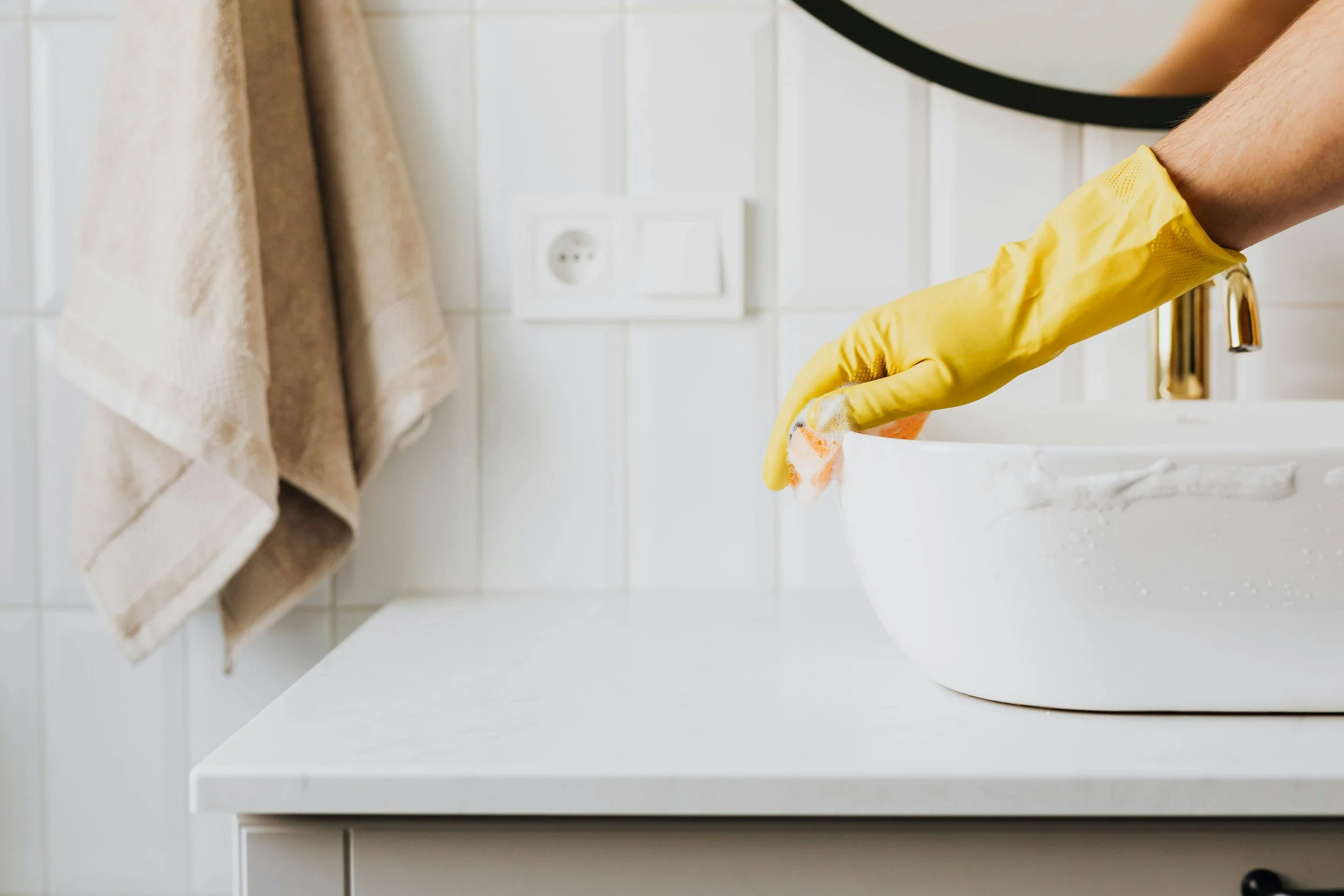 A person wearing yellow rubber gloves is washing a white bowl in a kitchen sink.