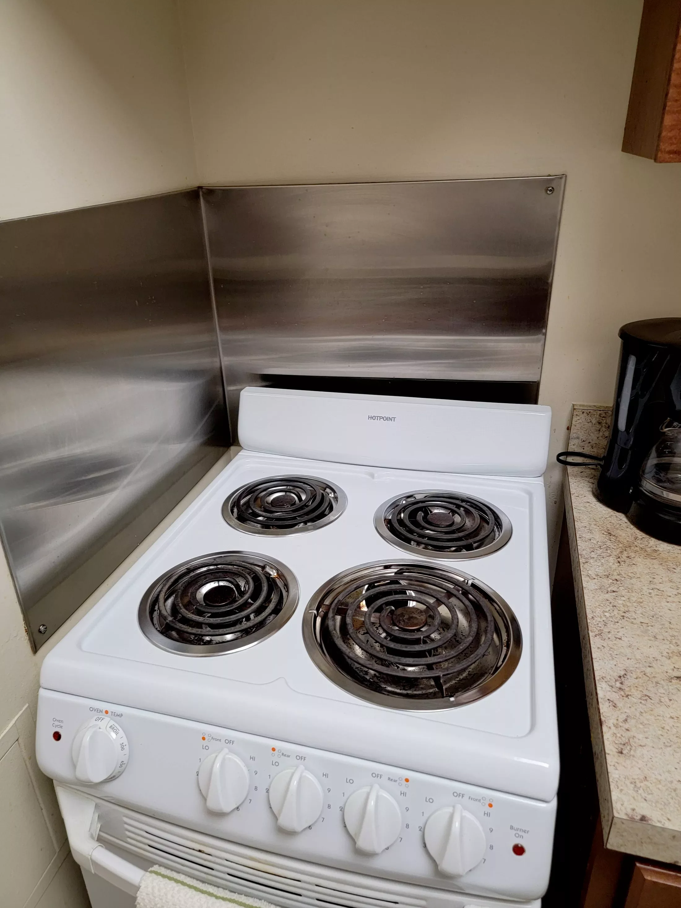 White electric stove with four coil burners, control knobs on the front, and a stainless steel backsplash, located in a kitchen with a granite countertop to the right.
