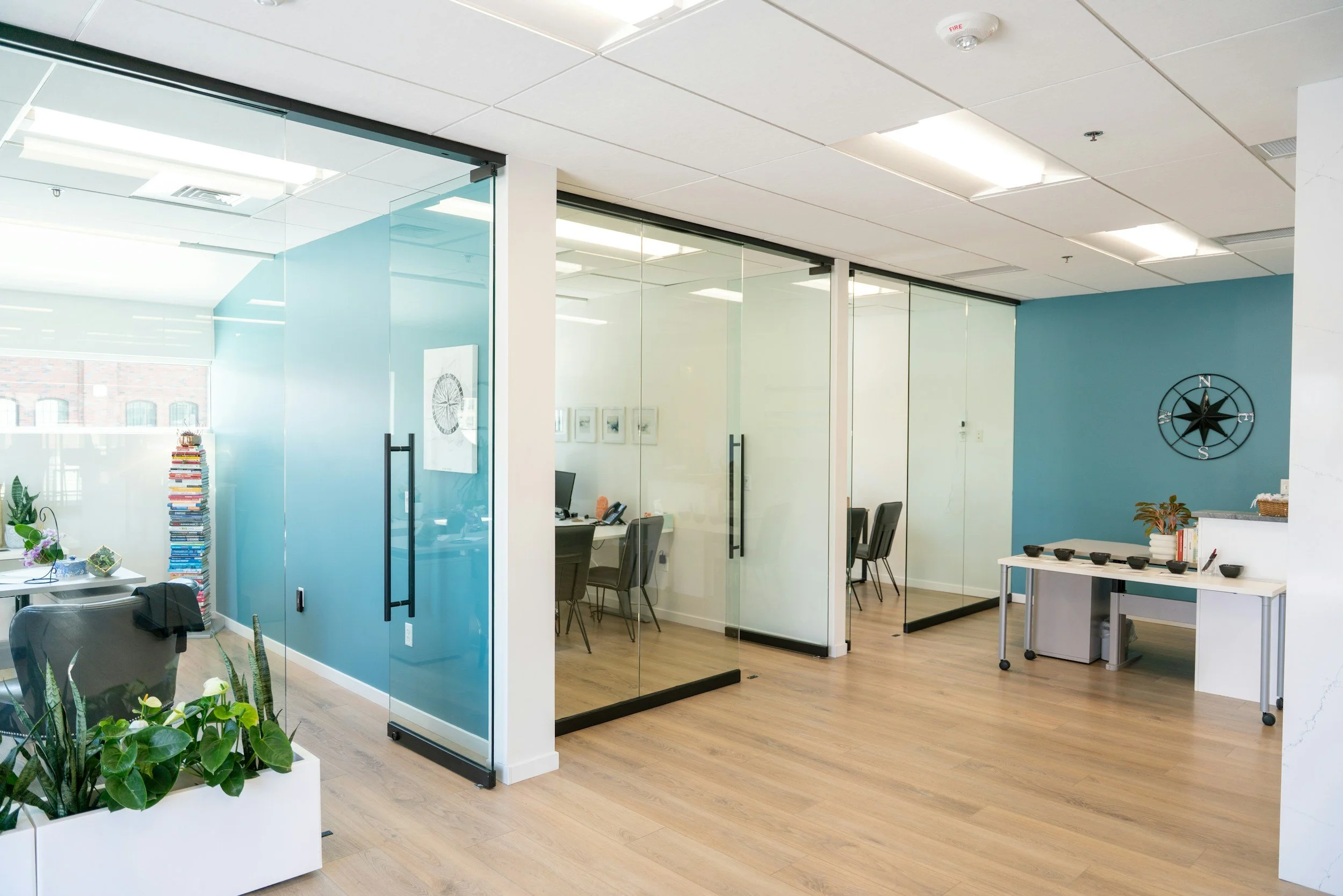 Modern office space with glass conference rooms, desks, chairs, indoor plants, and decorative elements like a wall clock on a blue accent wall.