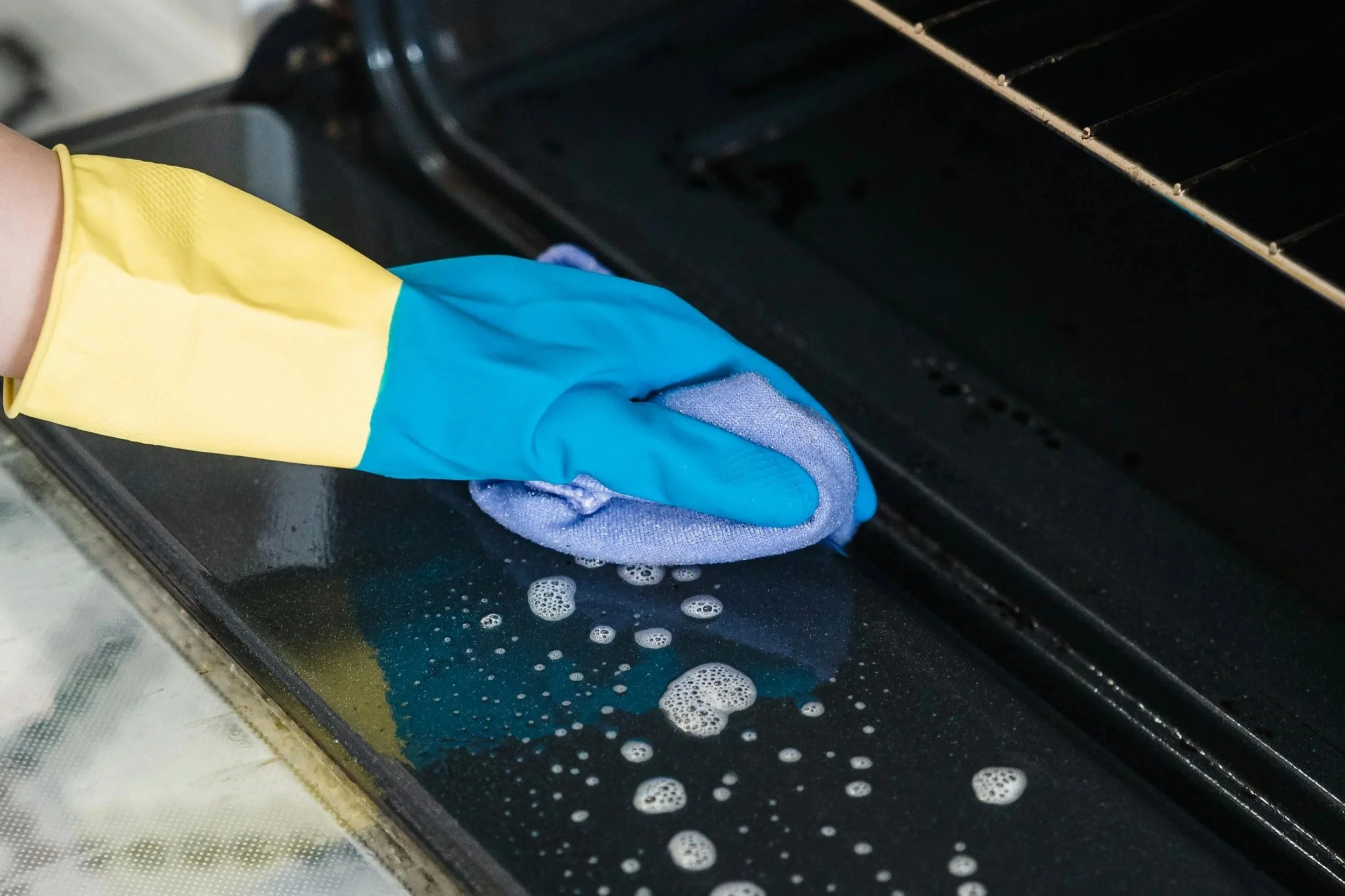 A person cleaning a black stovetop with a blue rubber glove and a purple cloth, creating soap suds.