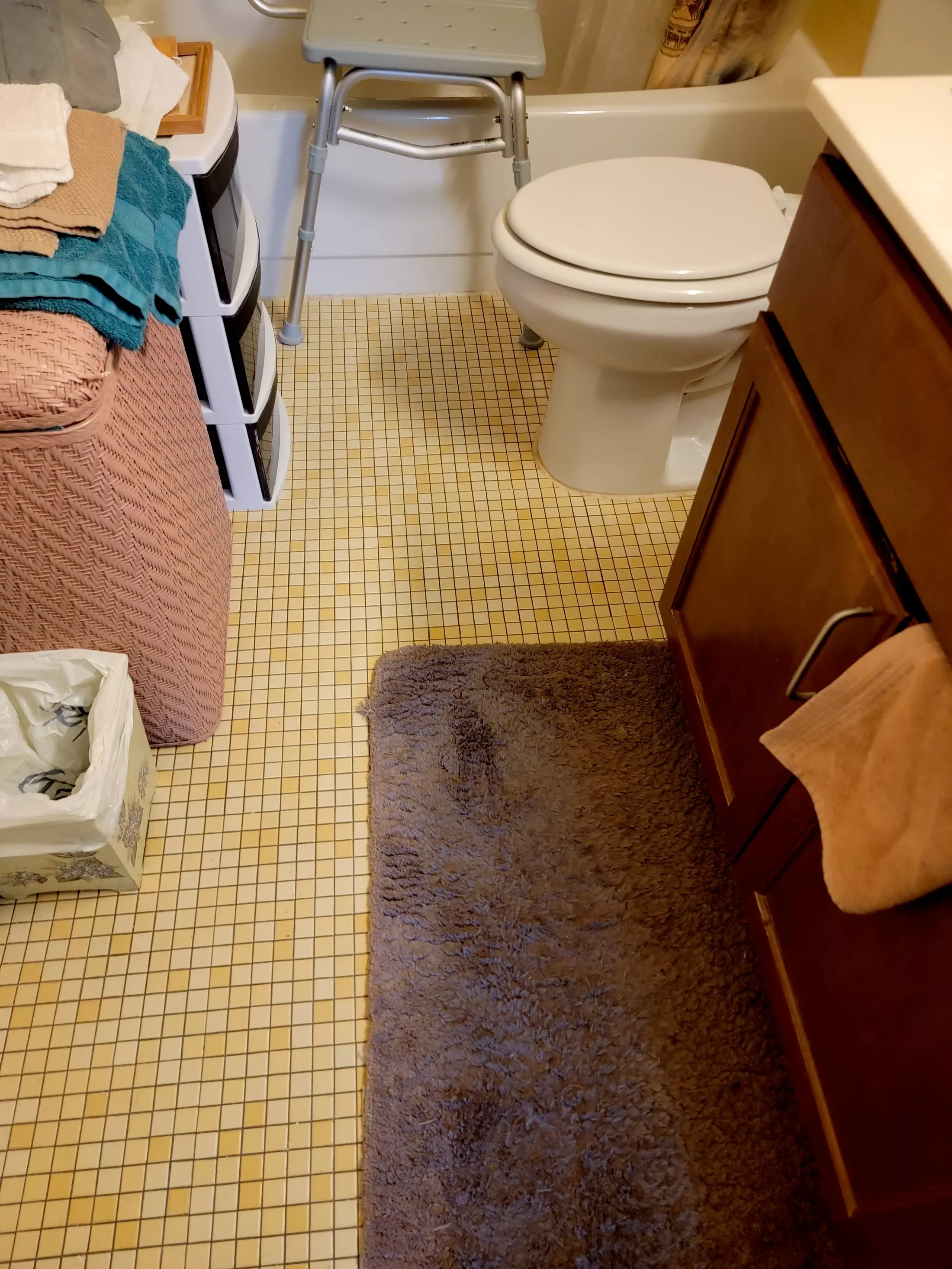 A bathroom with a toilet, a brown rug, a towel hanging on a cabinet door, a small plastic storage rack with towels, and a walker near the bathtub. The floor has small yellow tiles.