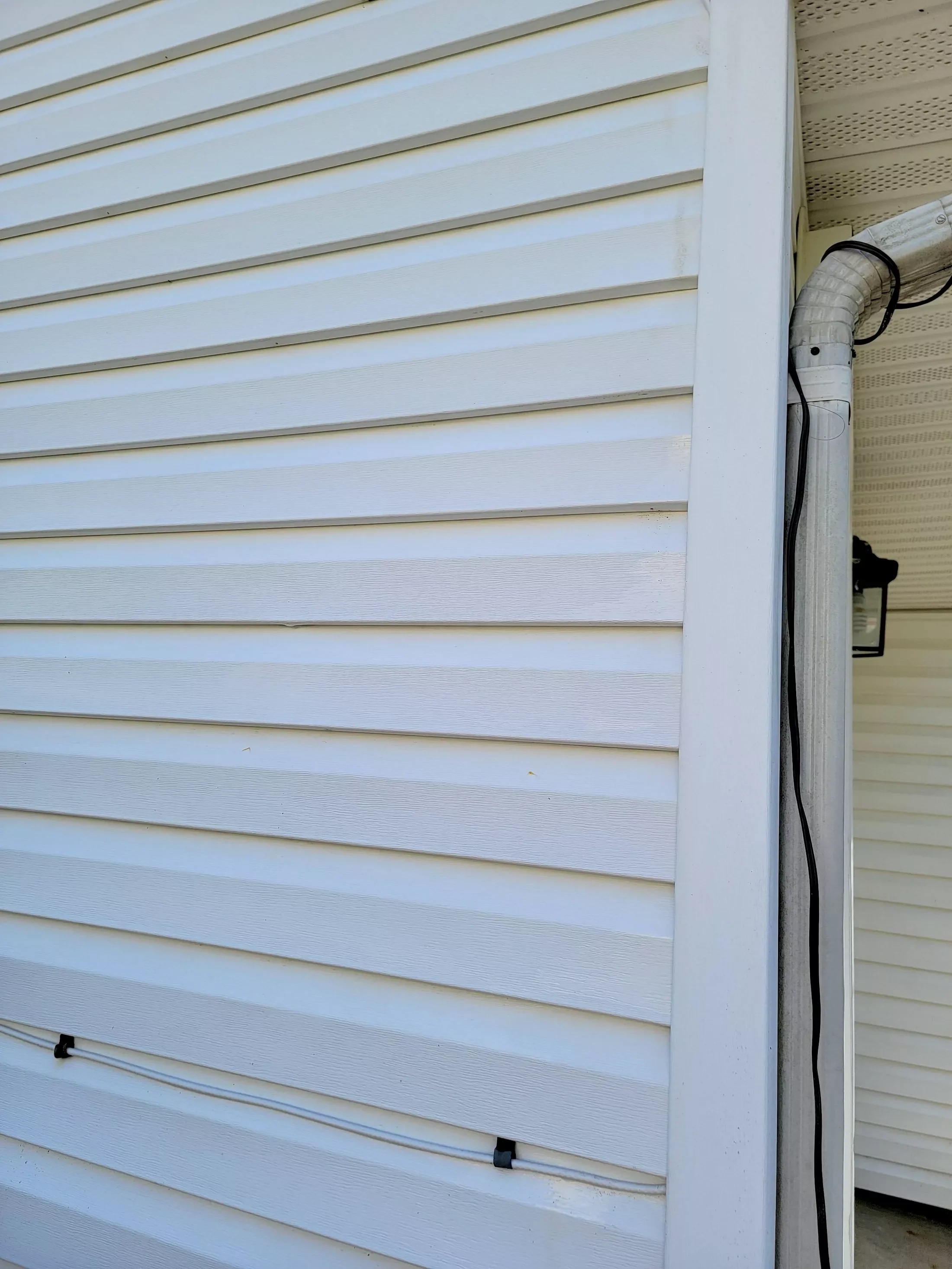 Close-up of white vinyl siding on the exterior of a house. A corner of the house's wall is visible with a partial view of a rain gutter and downspout, along with some black cables.