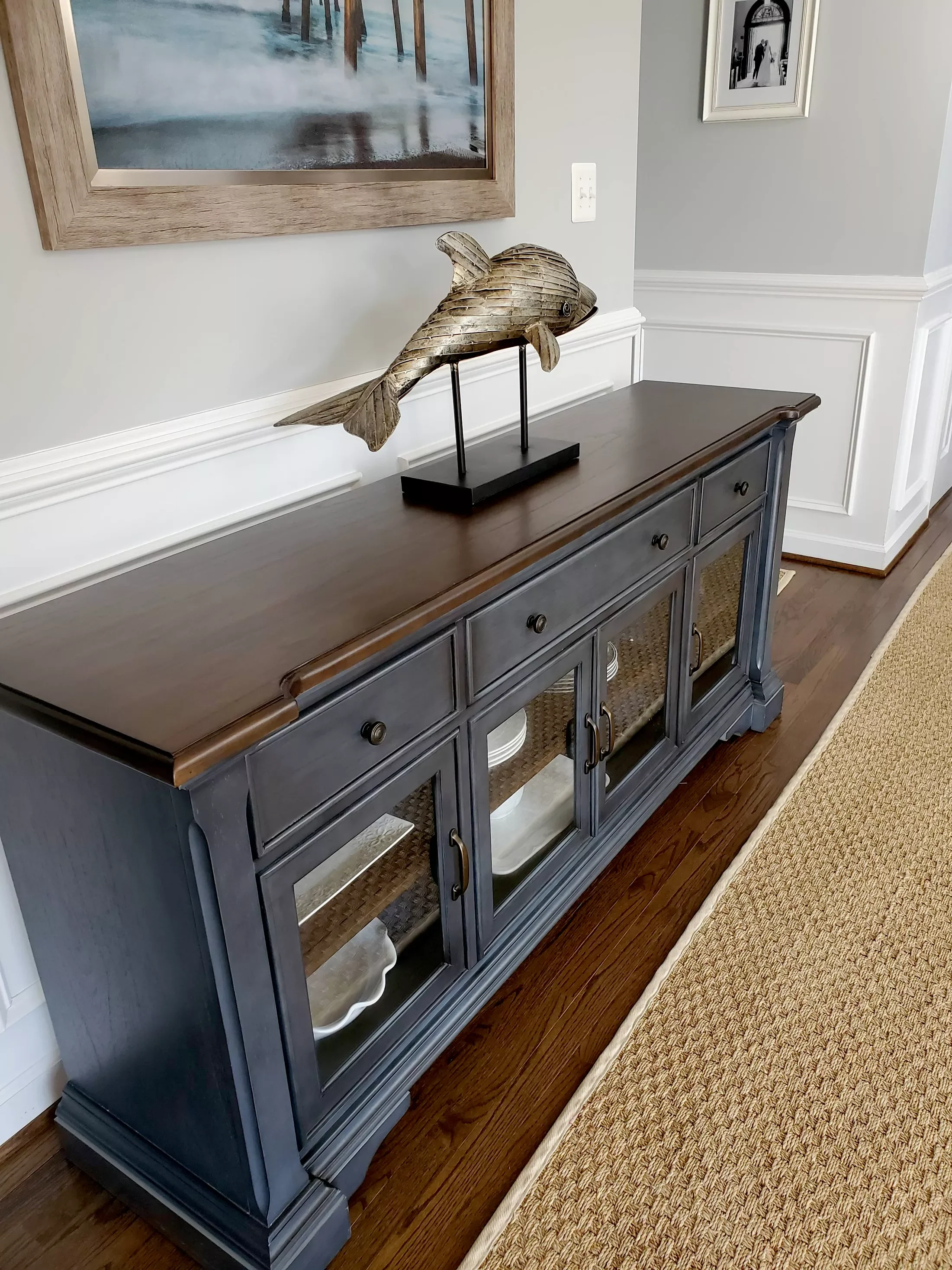 A black wooden sideboard with glass-paneled doors, situated on a hardwood floor with a beige carpet to the right. On top of the sideboard, there is a decorative wooden fish sculpture mounted on two vertical rods attached to a black rectangular base. 