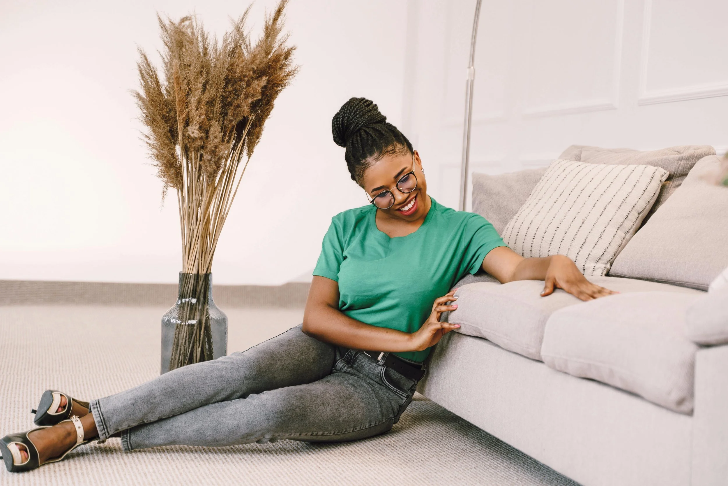 A woman with glasses and a bun hairstyle smiling and leaning on a light gray couch, with her arm resting on the sofa arm, in a well-lit room with a beige carpet, a tall vase with dried plants, and white walls.