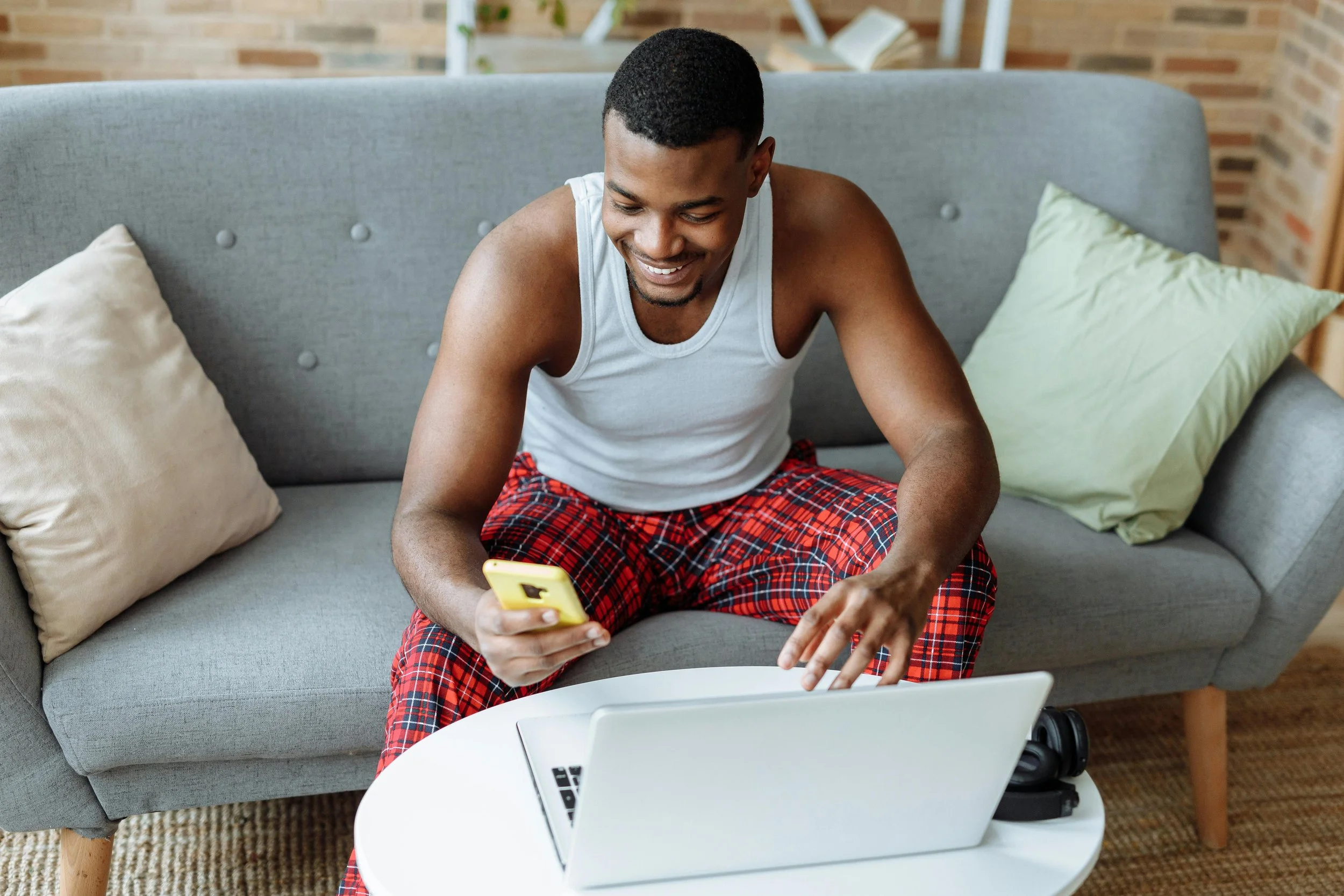 Young man in pajamas sitting on a couch, looking at his phone and smiling with a laptop on a table in front of him.