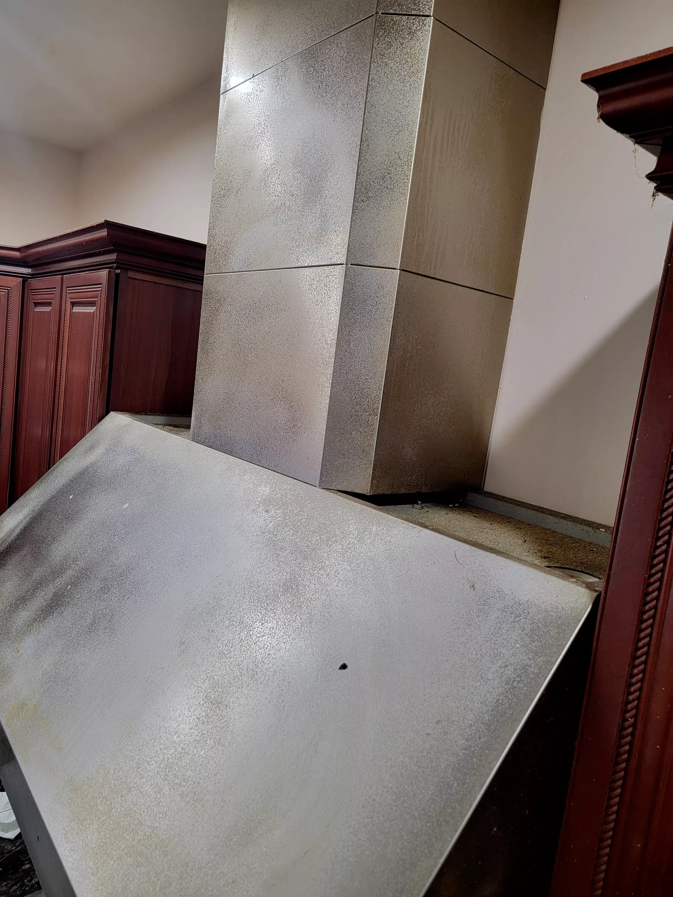 A partially dismantled stainless steel kitchen range hood with a detached cover in a kitchen with wooden cabinets.