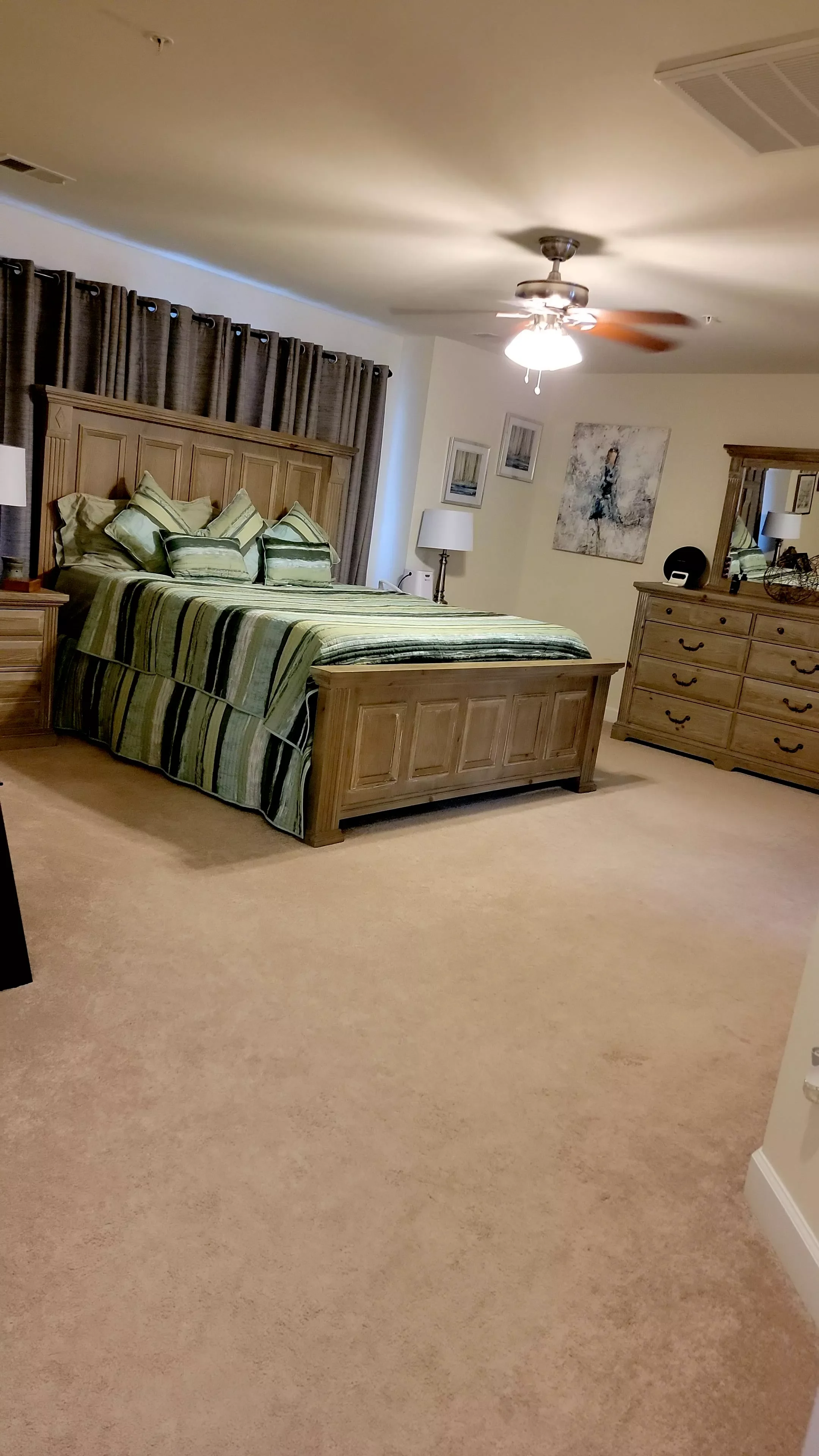 A bedroom with a large wooden bed, green striped bedding, and multiple pillows. There are wooden nightstands with lamps, a dresser with a mirror, and wall art. The room has beige carpet and a ceiling fan.