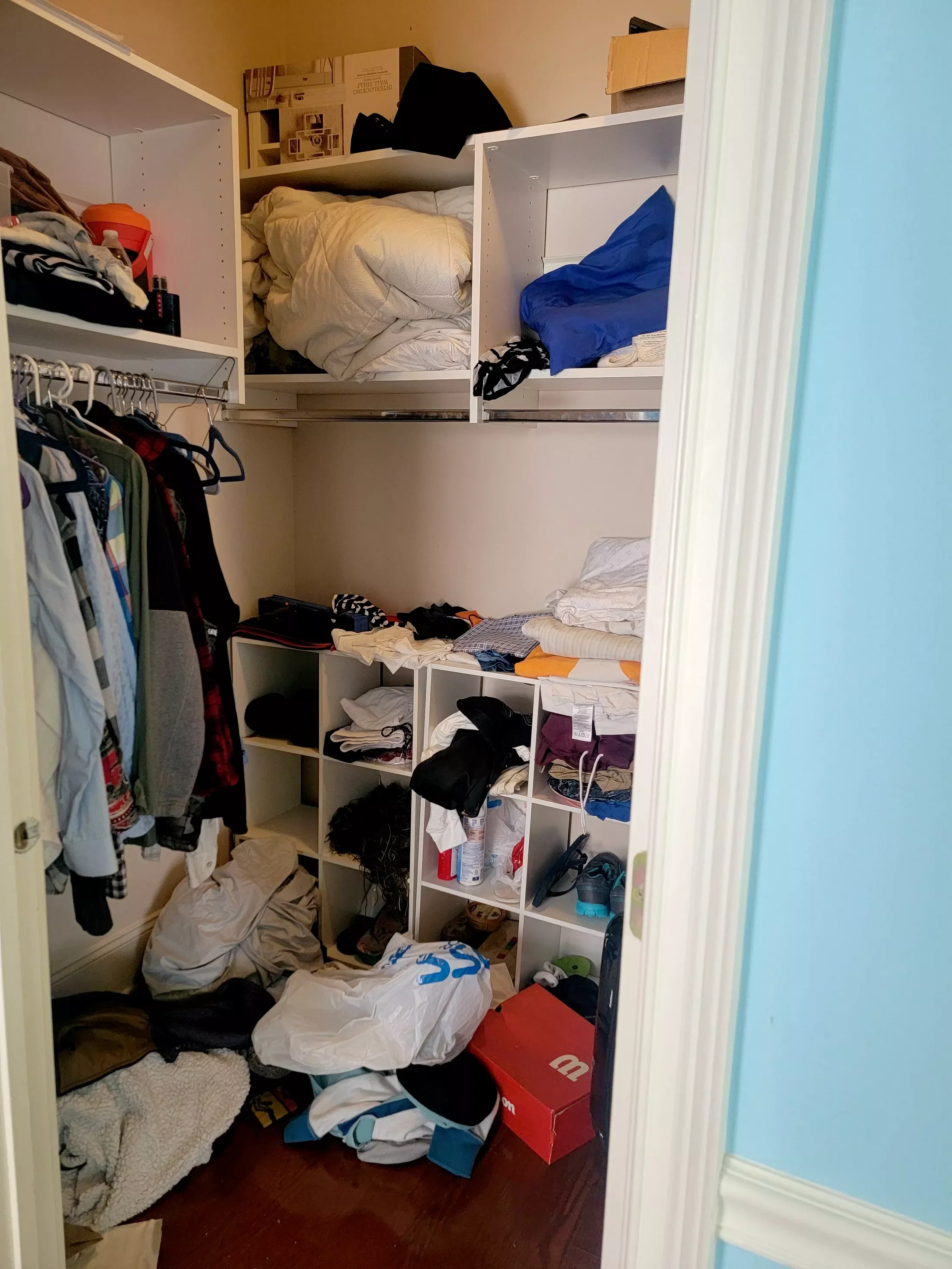 A cluttered walk-in closet with shelves and hanging clothes, with laundry on the floor and various items stored throughout the space.