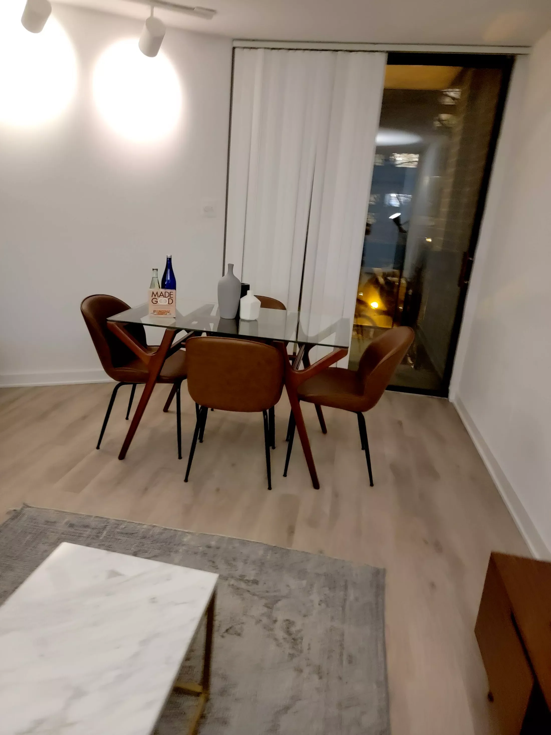 A dining area with a glass table and four brown chairs, decorative vases, and a window with vertical blinds overlooking a balcony at night.
