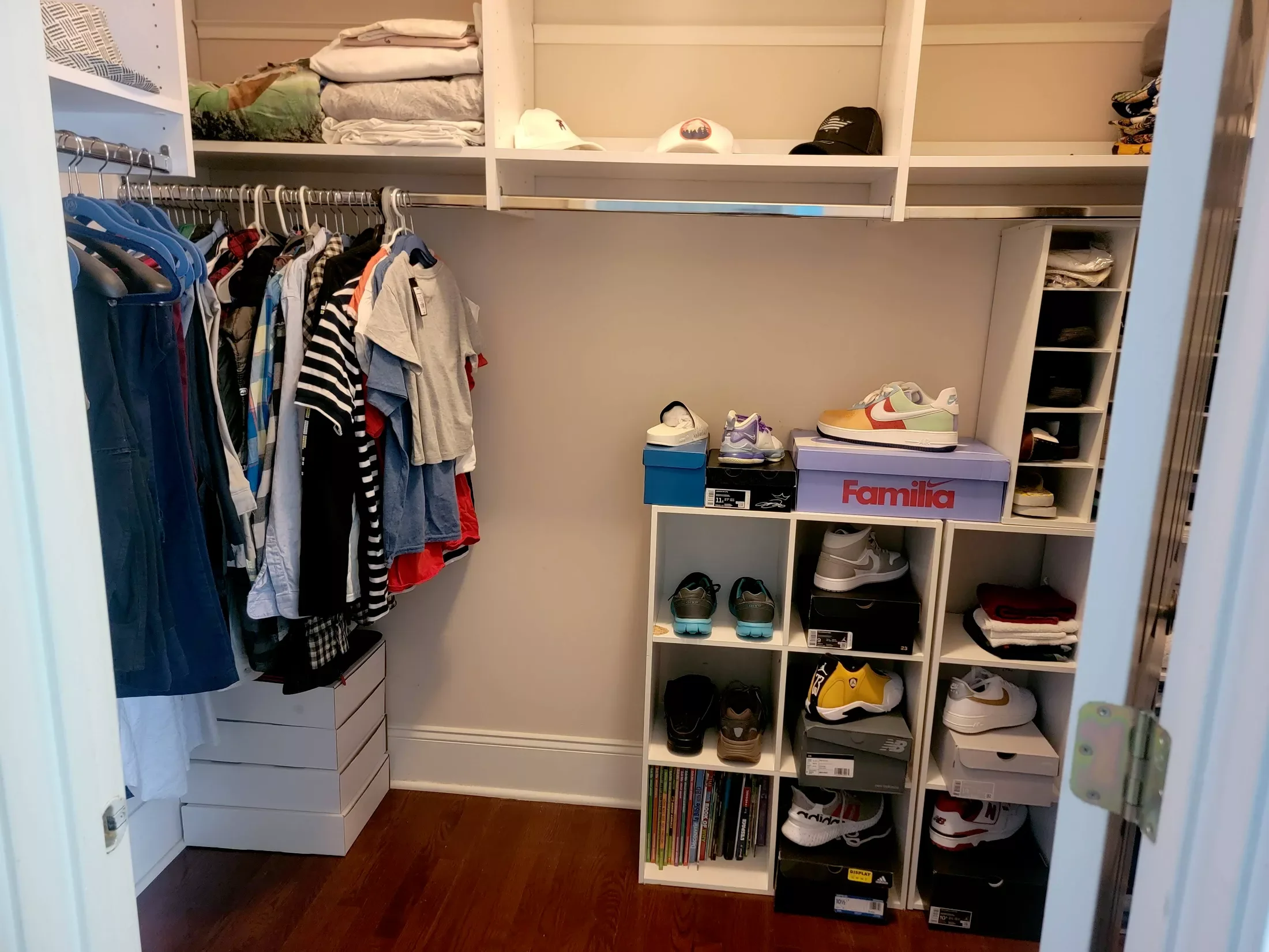 A walk-in closet with hanging clothes on the left, shelves and shoe boxes on the right, and a top shelf with folded clothes and hats.