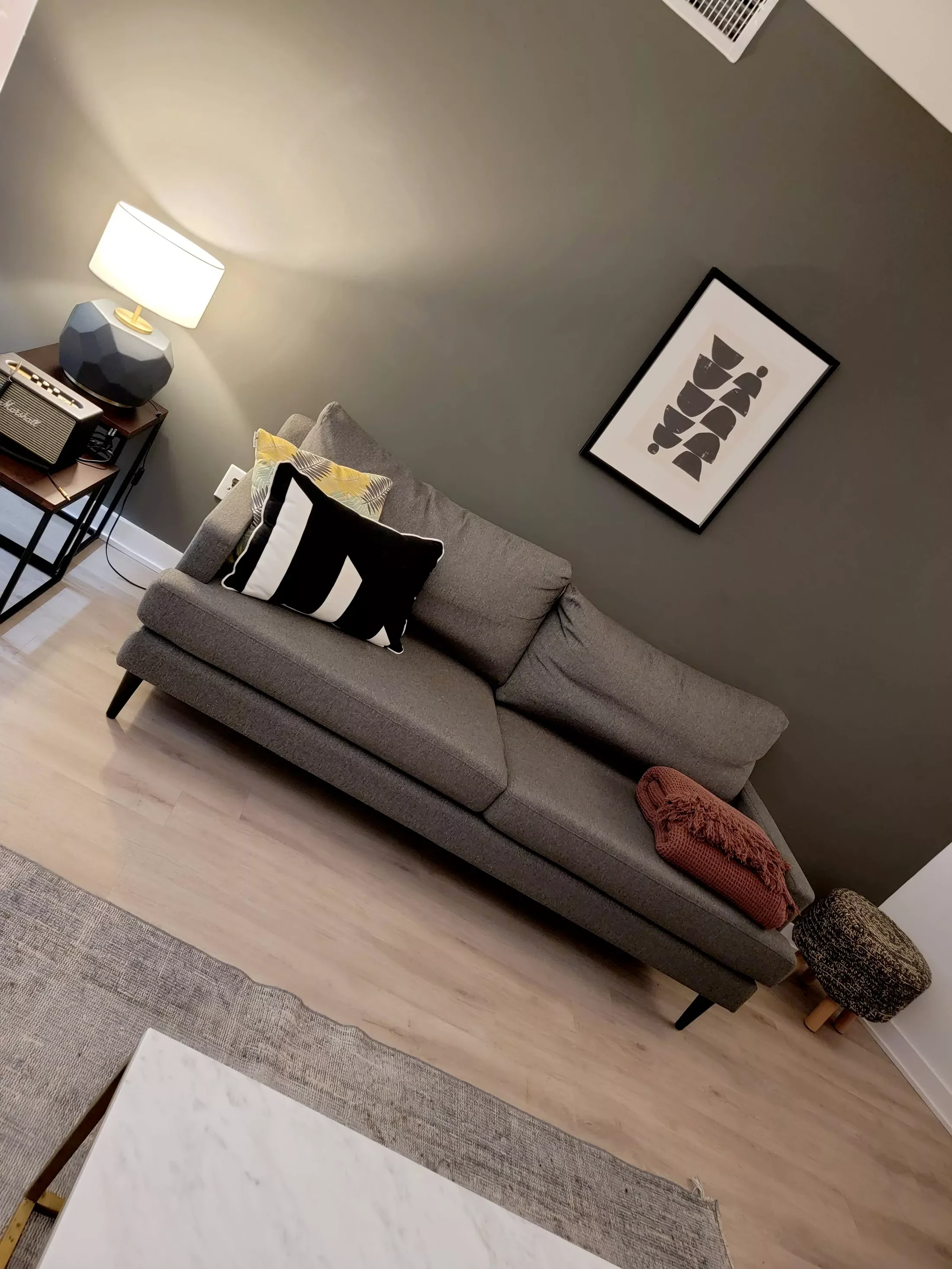 A modern living room with a gray sofa, black and white pillows, a red throw blanket on the sofa, a small side table with a lamp, decorative object, and an amplifier, a framed abstract art piece on the wall, and a small pouf on the floor.